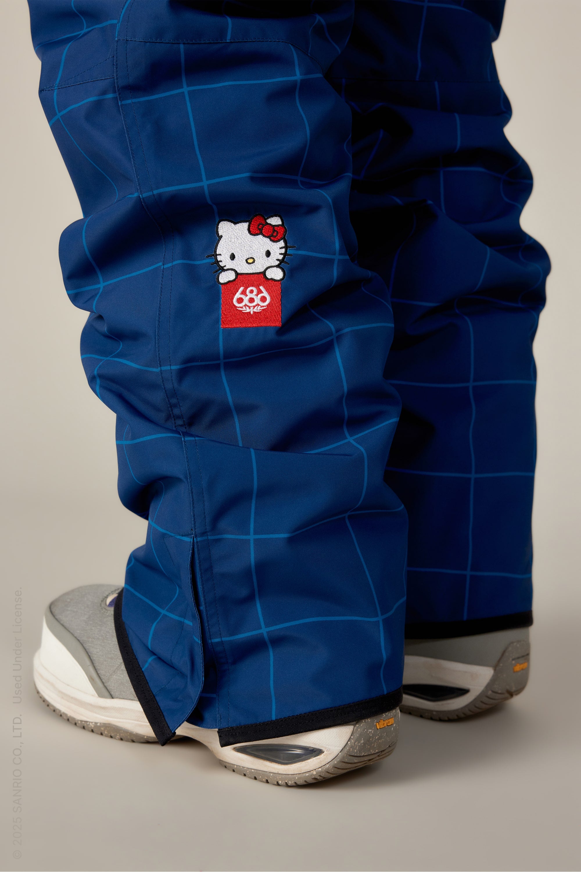 A close-up of someone wearing the 686 Women's Aura™ Insulated Cargo Pant in blue plaid with a small Hello Kitty patch and red 686 logo, paired with gray and white sneakers.