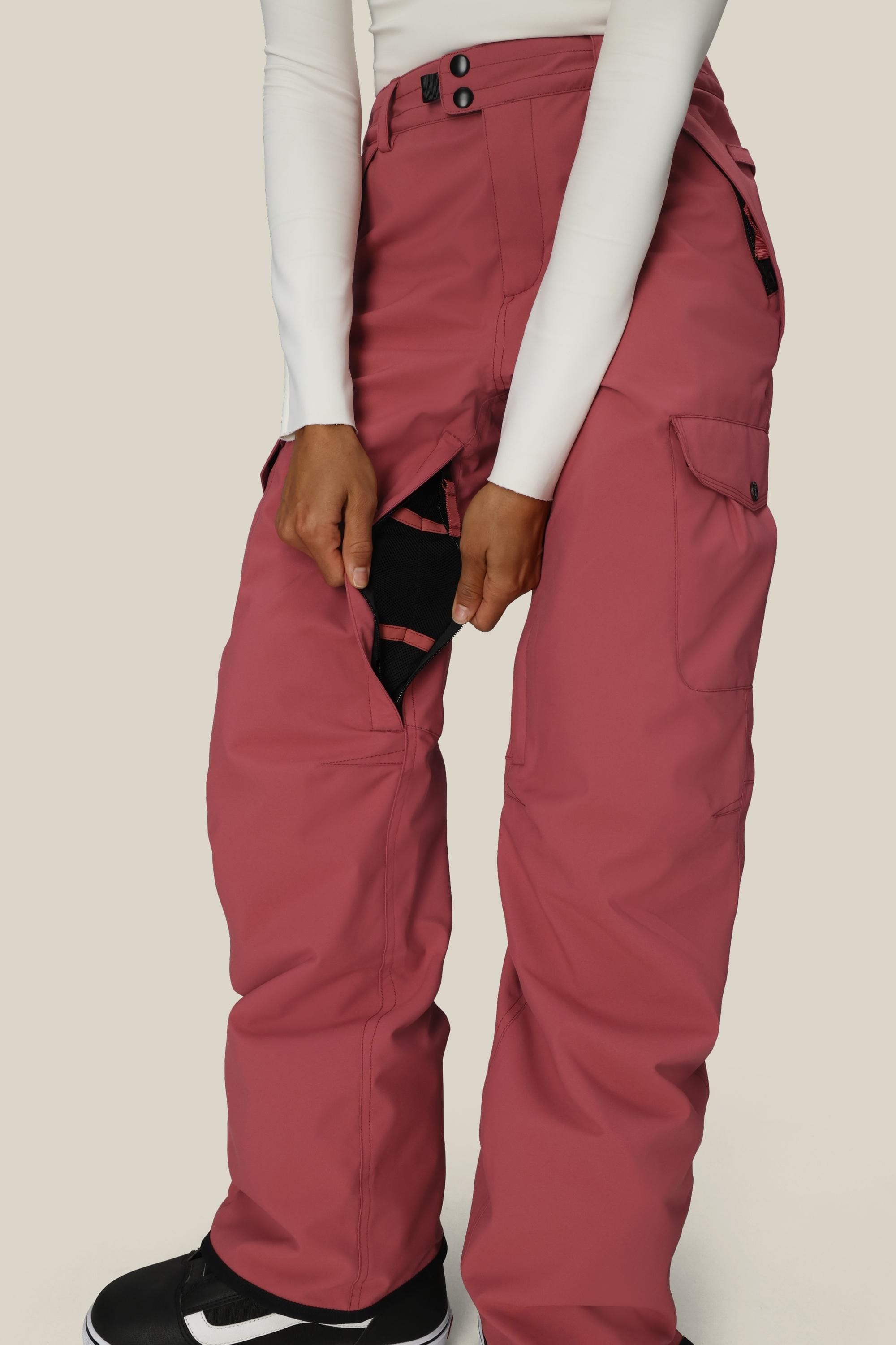 A person in rose-colored 686 Women's Aura™ Insulated Cargo Pant and a white long-sleeve top opens a black side vent to reveal the mesh lining, standing against a plain light background.