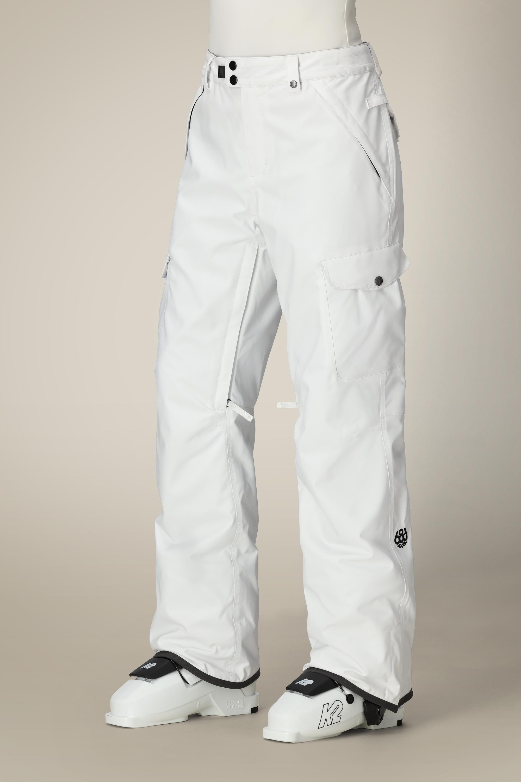A person wearing 686 Women's Aura™ Insulated Cargo Pant with black buttons and pockets stands in white ski boots labeled K against a plain beige background.