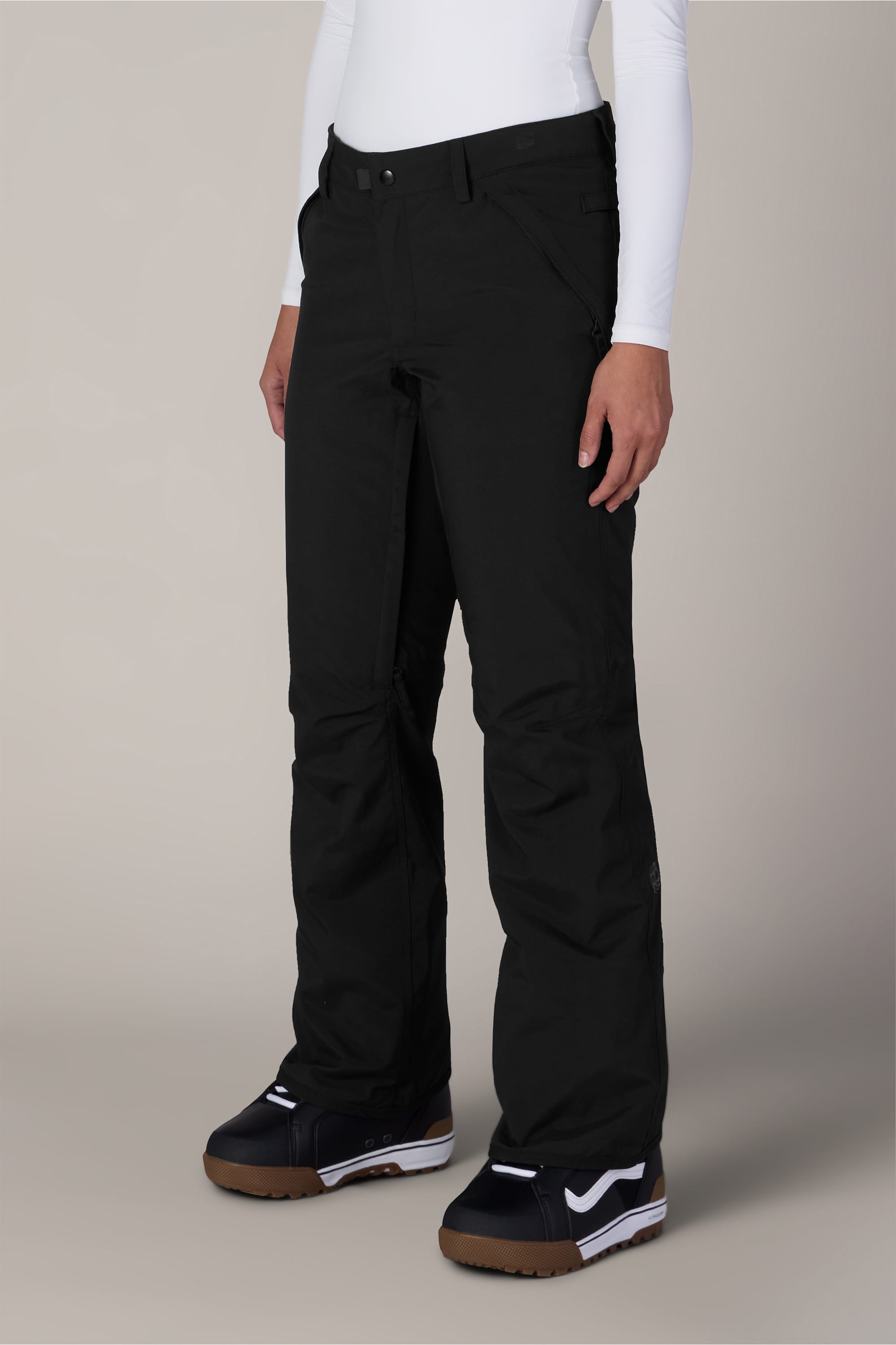 A person wearing the 686 Women’s Standard Shell Pant in black by 686, paired with black snow boots featuring white accents and a white long-sleeve top, stands against a plain beige background.
