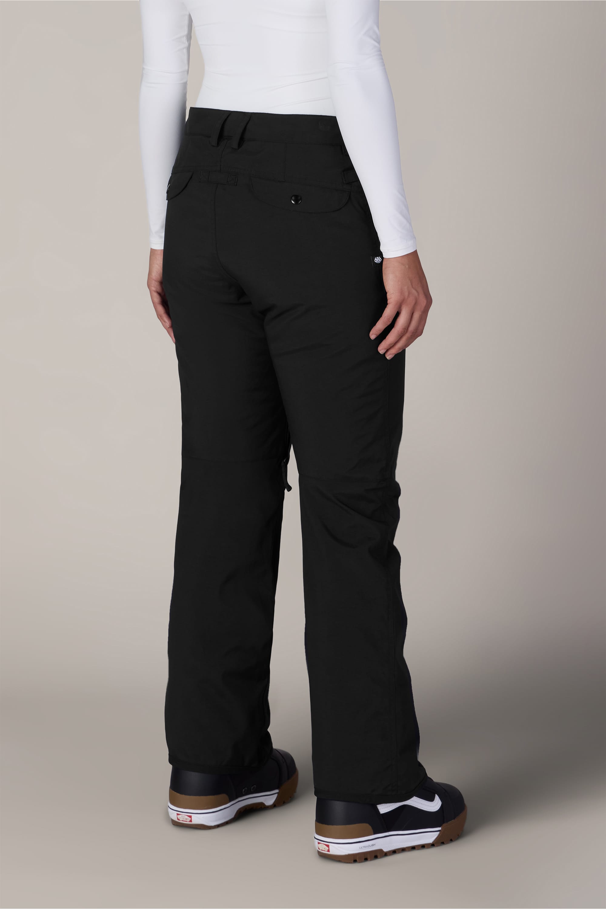 A person wearing the 686 Women’s Standard Shell Pant by 686, paired with a white long-sleeve shirt and black sneakers with white soles, is standing with their back to the camera against a plain background.