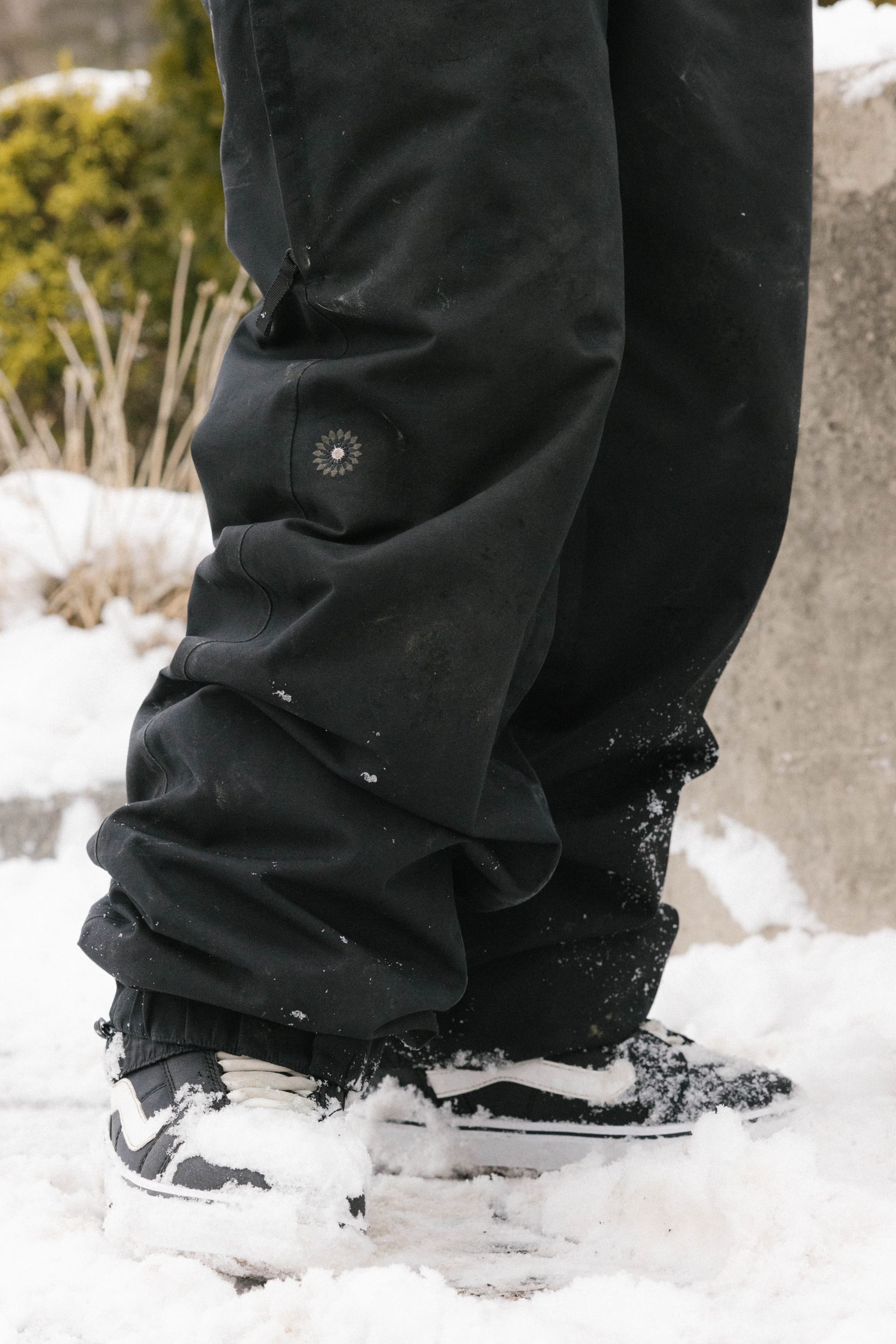Wearing 686 Women's Outline™ Shell Pant and black-and-white sneakers, a person stands on snow with patches of it on their shoes and pants; greenery appears blurred in the background.
