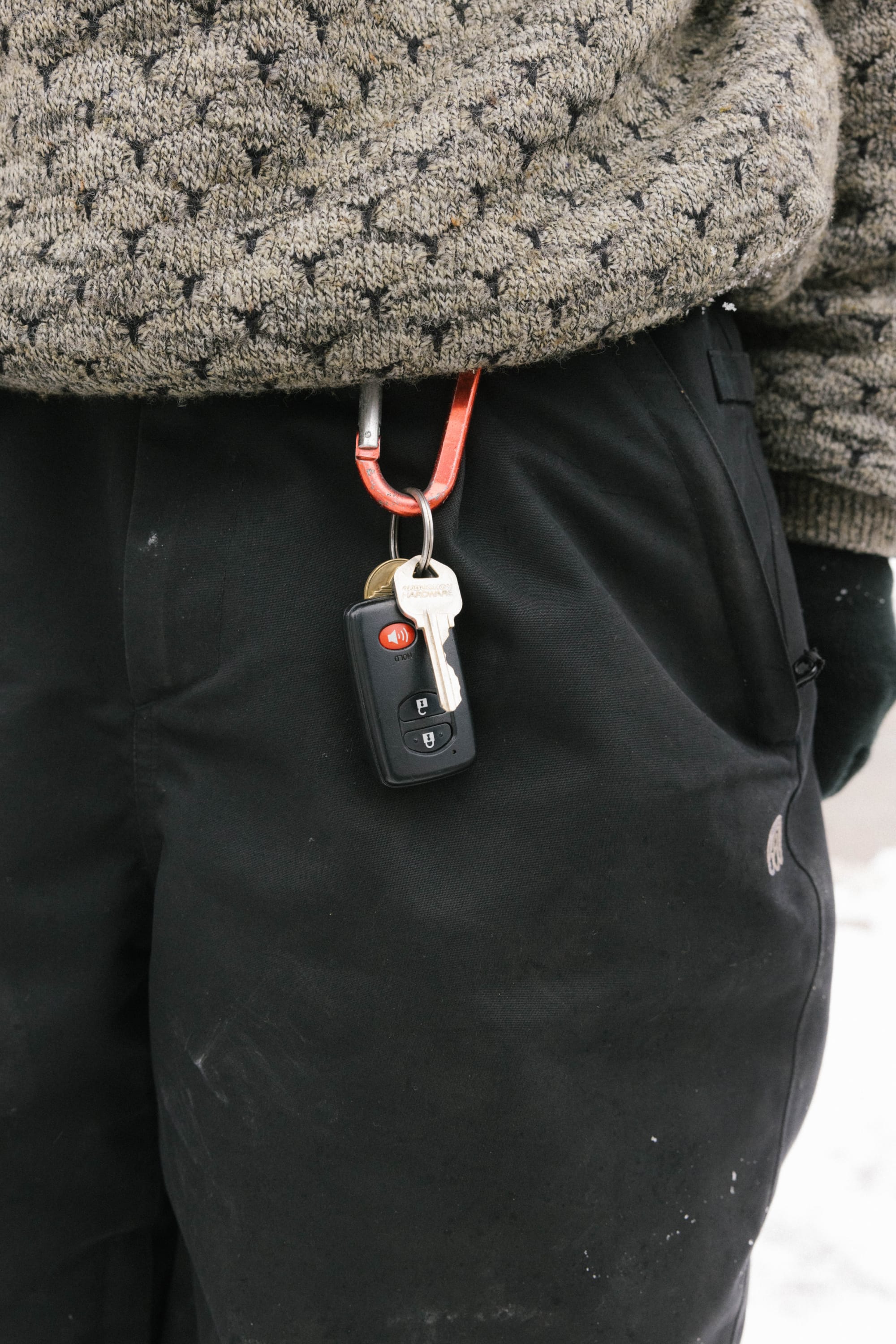 A person wearing 686 Women's Outline™ Shell Pant and a patterned sweater has car keys and a remote on a red carabiner attached to a belt loop. Snow covers the ground in the background.