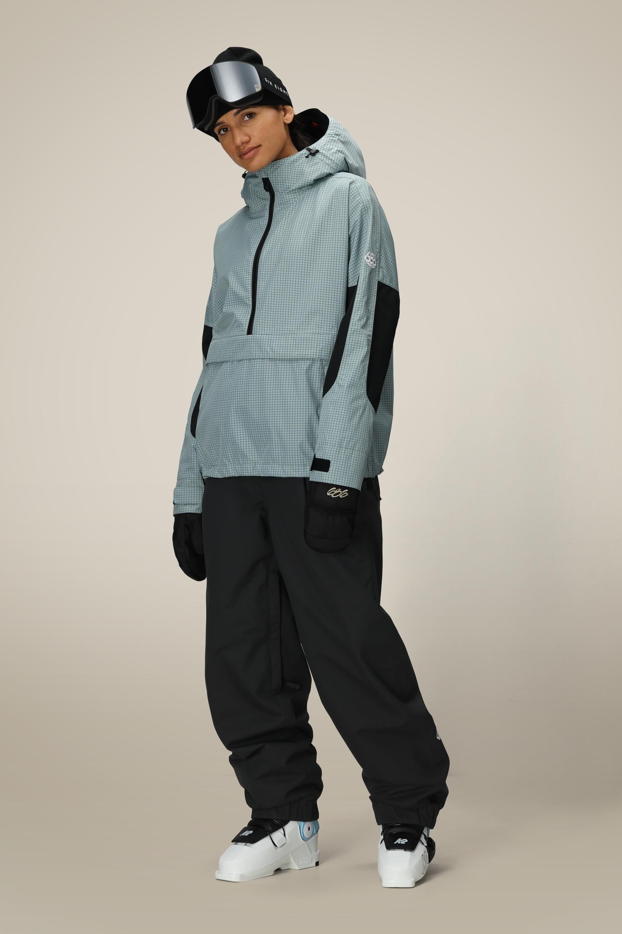 A person stands against a plain background wearing a light gray hooded jacket, black gloves, white snow boots, ski goggles on their head, and 686 Women's Outline™ Shell Pant by 686.