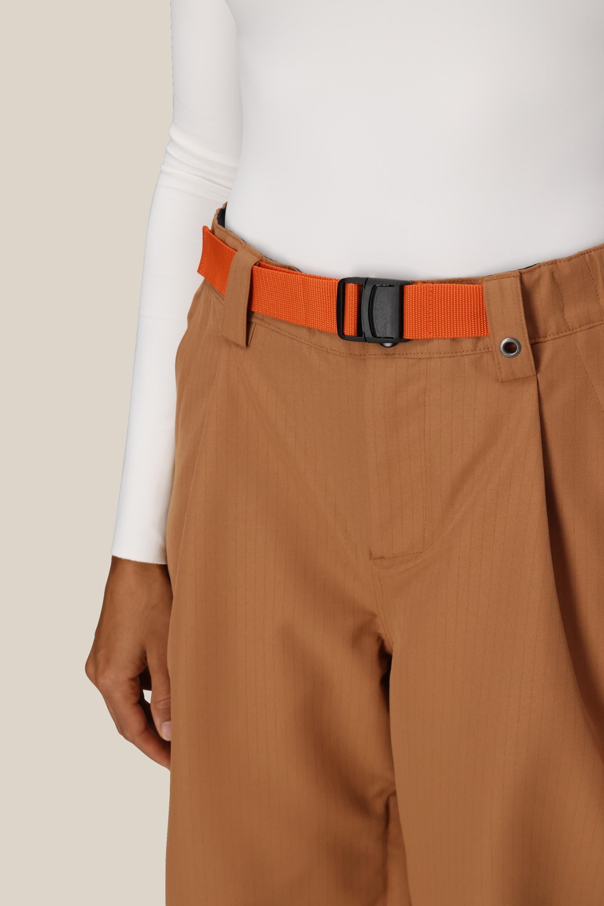 A person stands against a plain beige background, wearing the 686 Women's Outline™ Shell Pant with an orange belt and a white long-sleeve top.