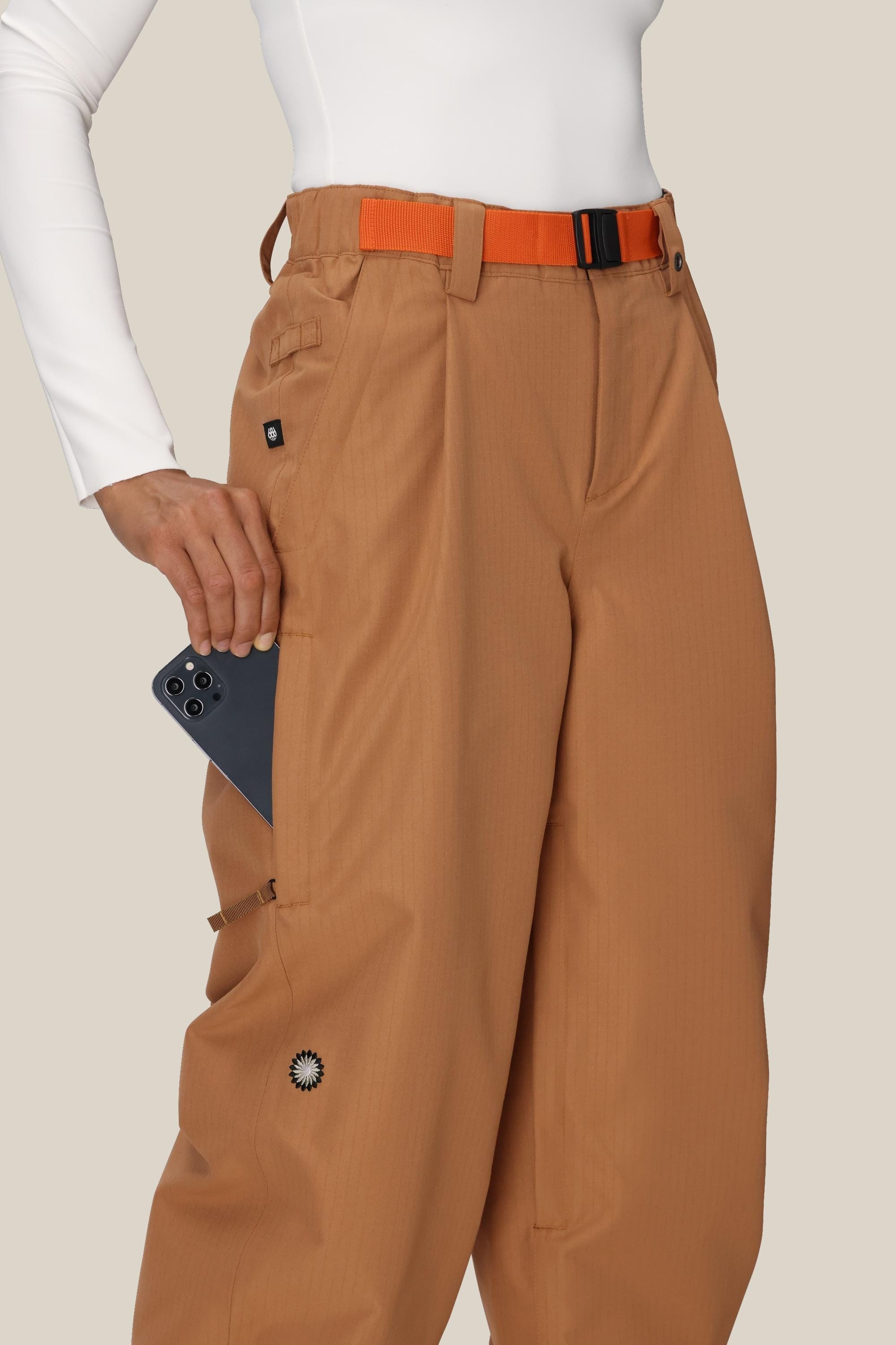 A person wears 686 Women's Outline™ Shell Pant by 686 in tan-brown with a wide-leg fit and large side pocket, styled with an orange belt and white long-sleeve top, placing a black smartphone into the pocket against a plain light background.