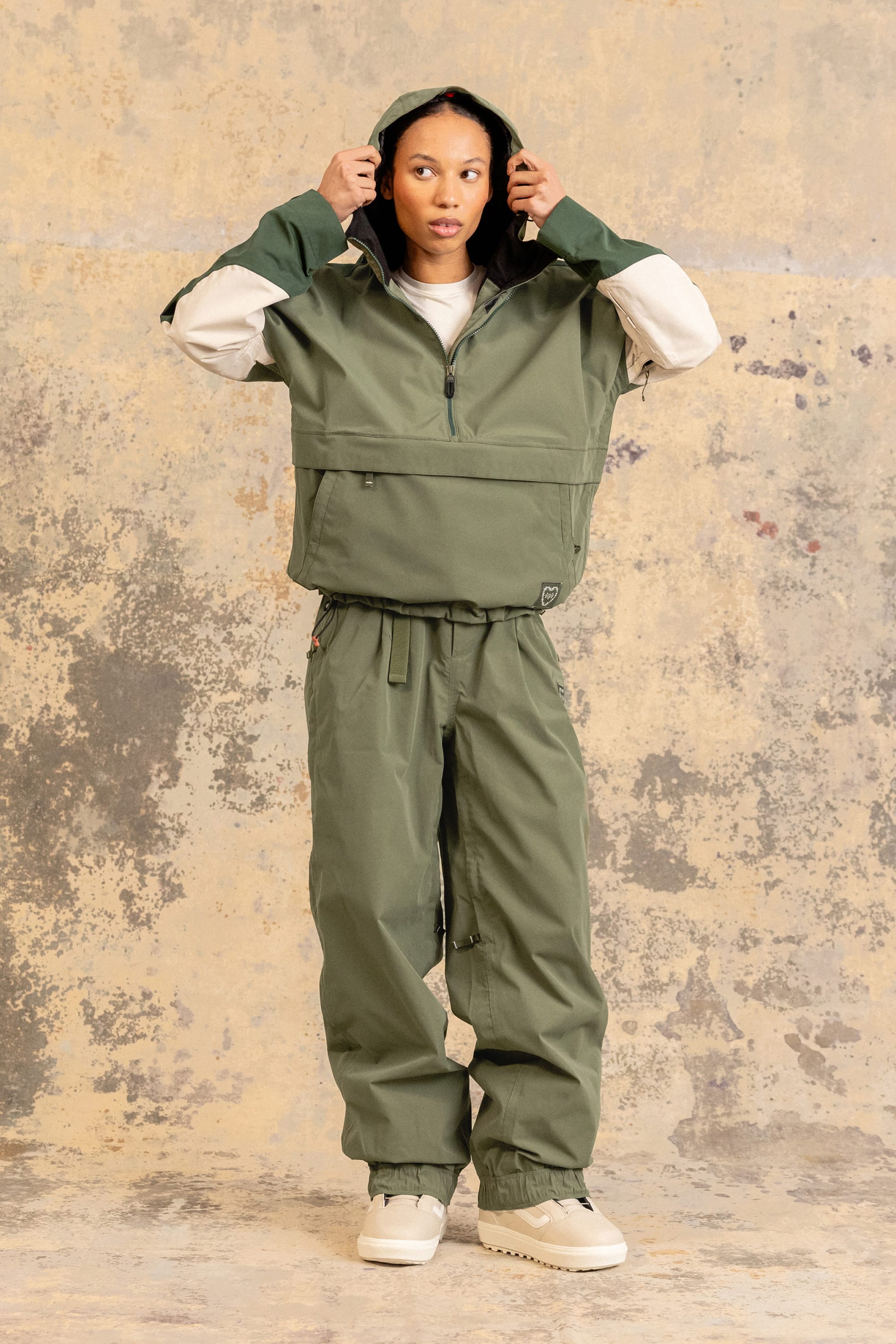 A person stands facing forward, wearing an olive green hooded jacket and the 686 Women’s Outline™ Shell Pant by 686, paired with beige shoes, against a textured beige and gray background.