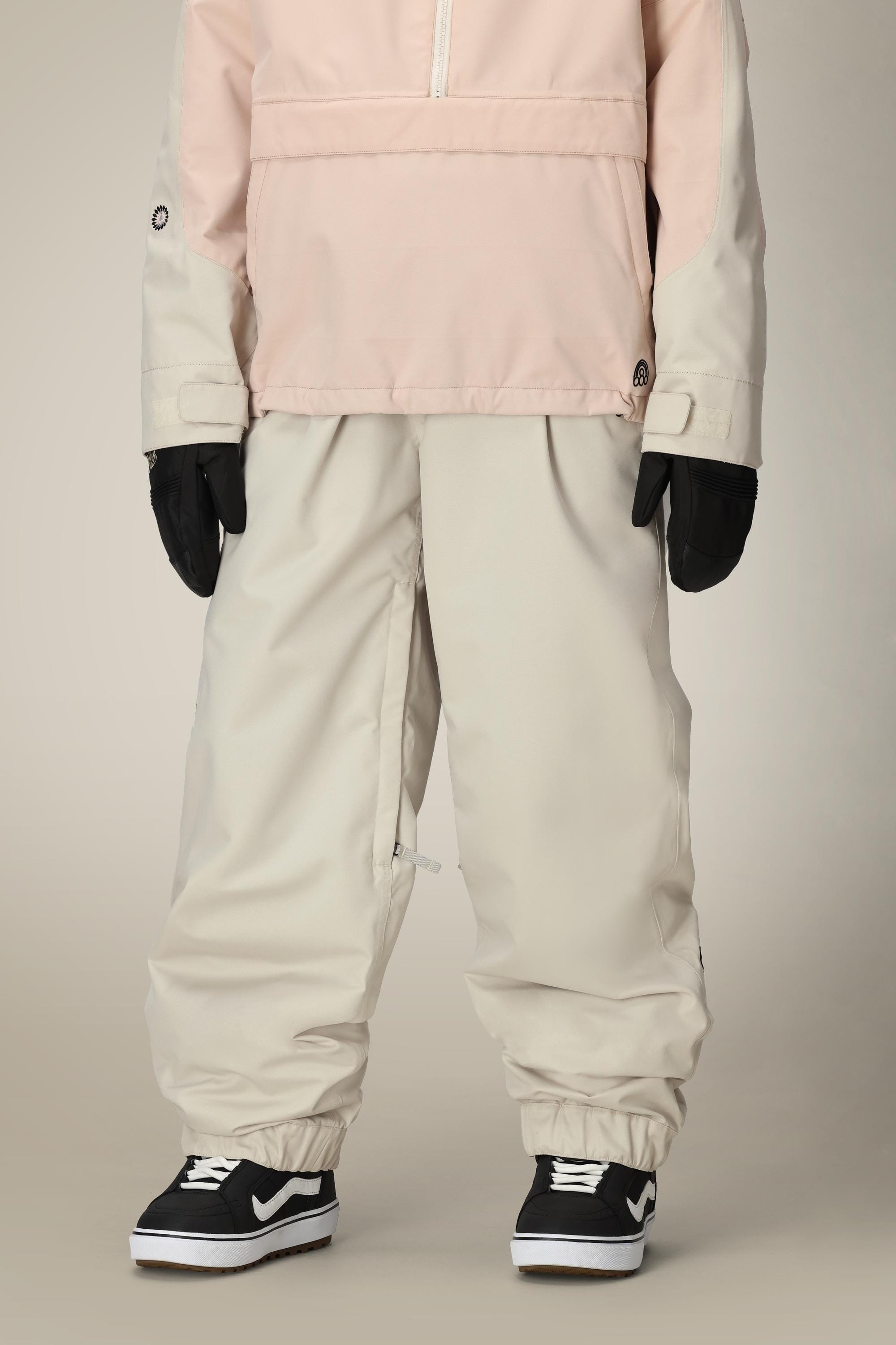 A person wears 686 Women's Outline™ Shell Pant in light pink and cream, paired with black gloves and black-and-white Vans, standing against a plain background. The focus is on the stylish waterproof polyester pants by 686.