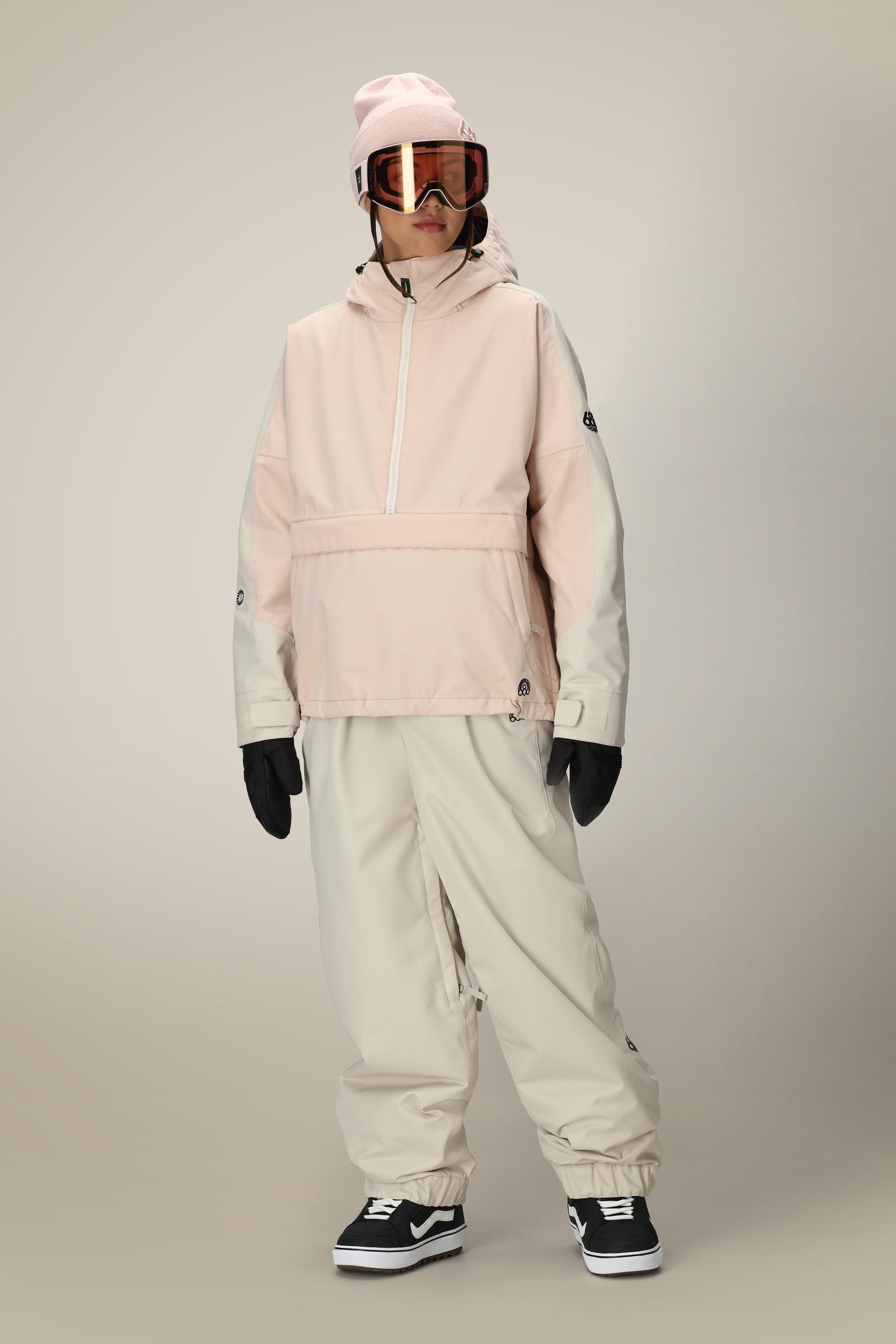 A person in a light pink hooded jacket, matching beanie, 686 Women's Outline™ Shell Pant by 686, black gloves, ski goggles, and black Vans sneakers stands against a plain, light background.