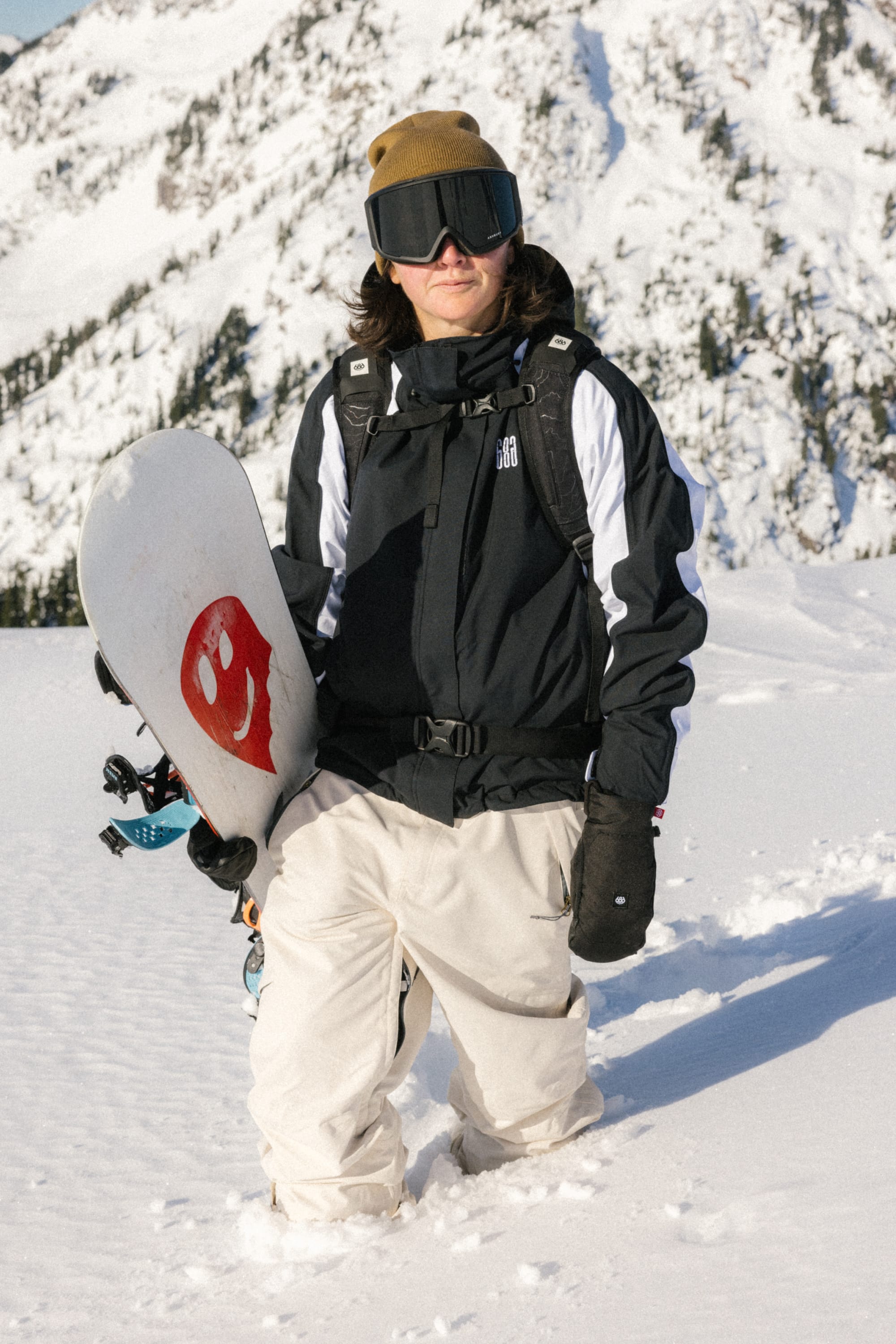 A snowboarder in a black jacket, 686 Women's Outline™ Shell Pant, brown beanie, and goggles stands in the snow holding a red snowboard, with snow-covered mountains in the background.