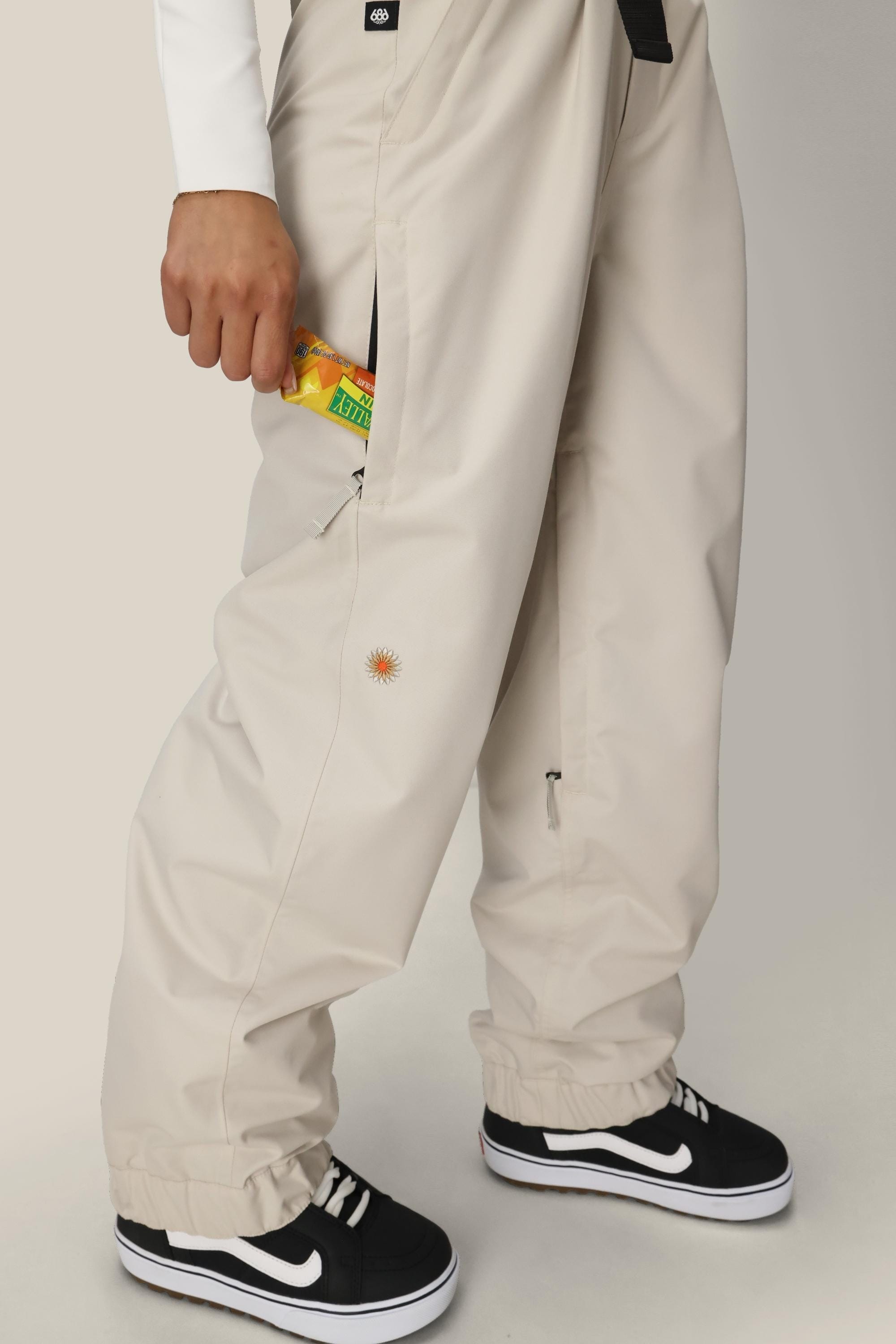 A person wearing 686 Women's Outline™ Shell Pant stands holding a small pack of Lemonhead candy in their right hand, partially tucked into the pants pocket, paired with black Vans sneakers.
