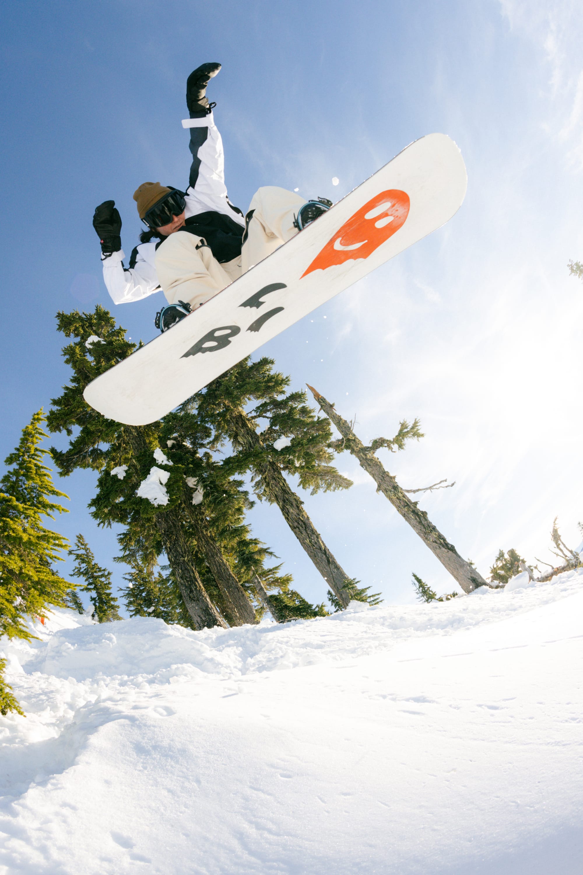 A snowboarder soars above snowy ground, framed by evergreens and blue sky, performing a trick in 686 Women's Outline™ Shell Pant. Her snowboard sports bold orange and black graphics.