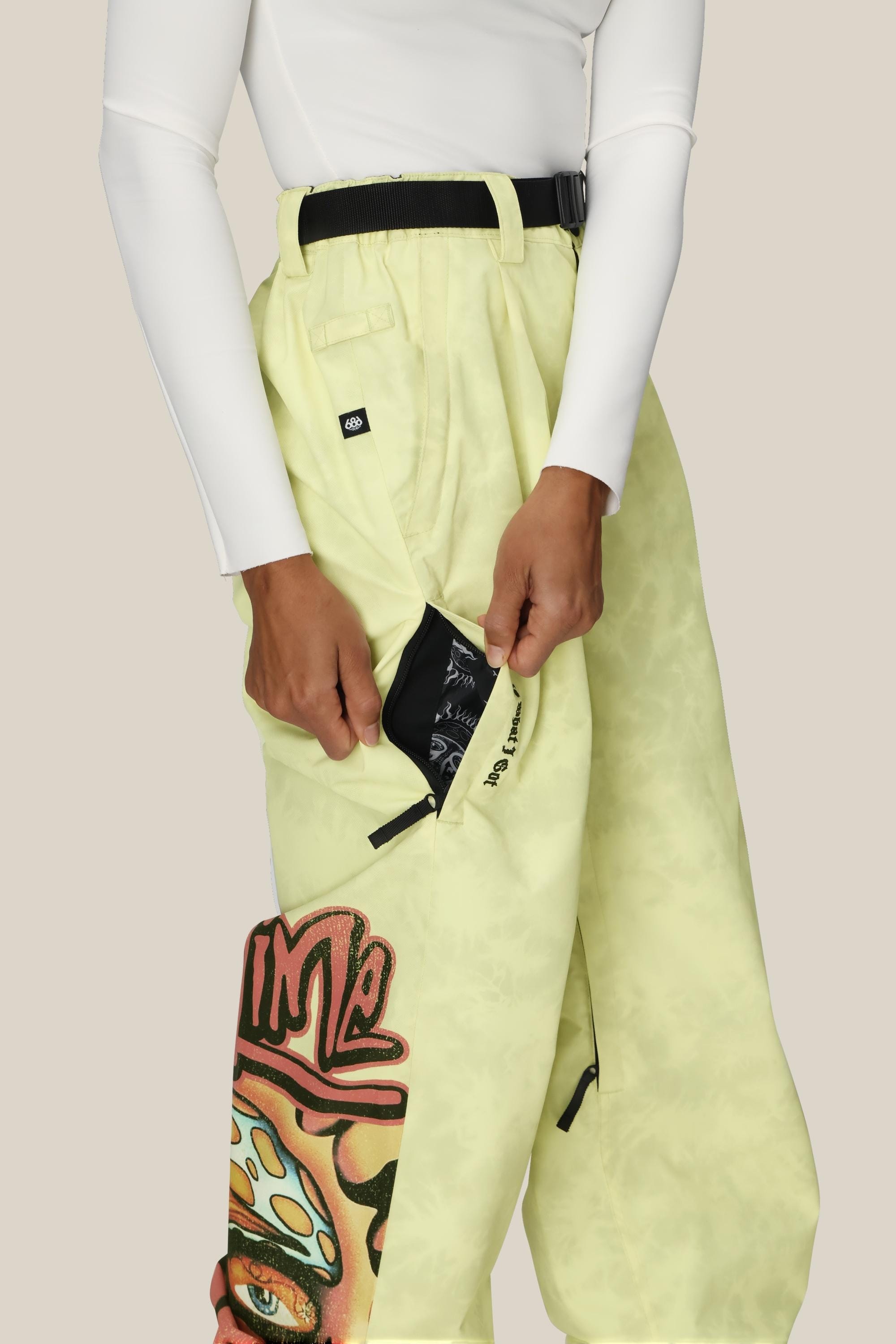 A woman wearing the 686 Womens Sublime Shell Pant in pale yellow with a colorful graphic and a white long-sleeve top unzips a large side pocket, revealing its black inner lining.