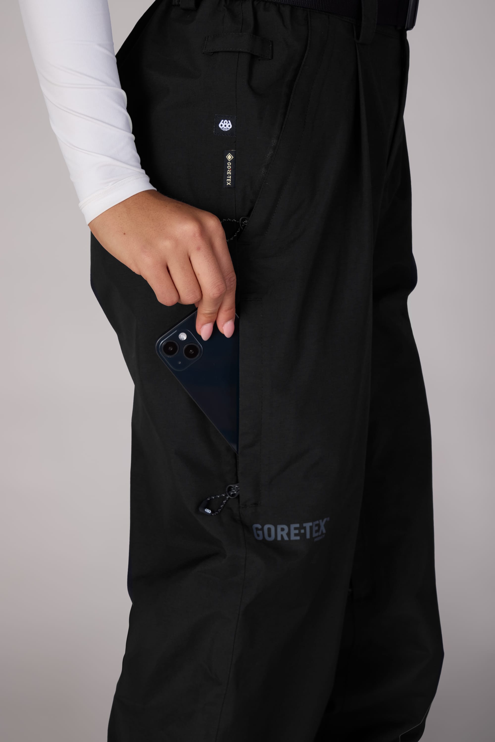 A person with light skin in a long-sleeve white shirt puts a black smartphone into the side zipper pocket of 686 Women's GORE-TEX Outline™ Shell Pant, made with waterproof woven fabric.