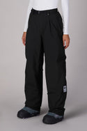 A person wears 686 Women's GORE-TEX Outline™ Shell Pant by 686—loose-fitting black waterproof pants with a belt and ankle patch—paired with a white long-sleeve top and black snow boots, standing on a neutral background.