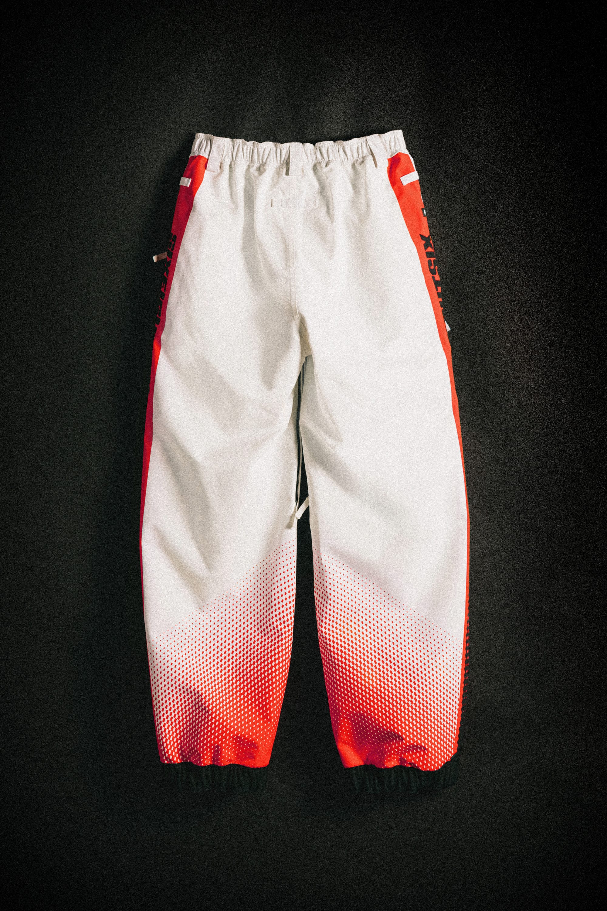 The 686 Women's Fox Racing Shell Pant by 686 features white fabric with red side panels, a gradient red dot pattern near the cuffs, and black elastic bands at the ankles, shown laid flat against a dark background.