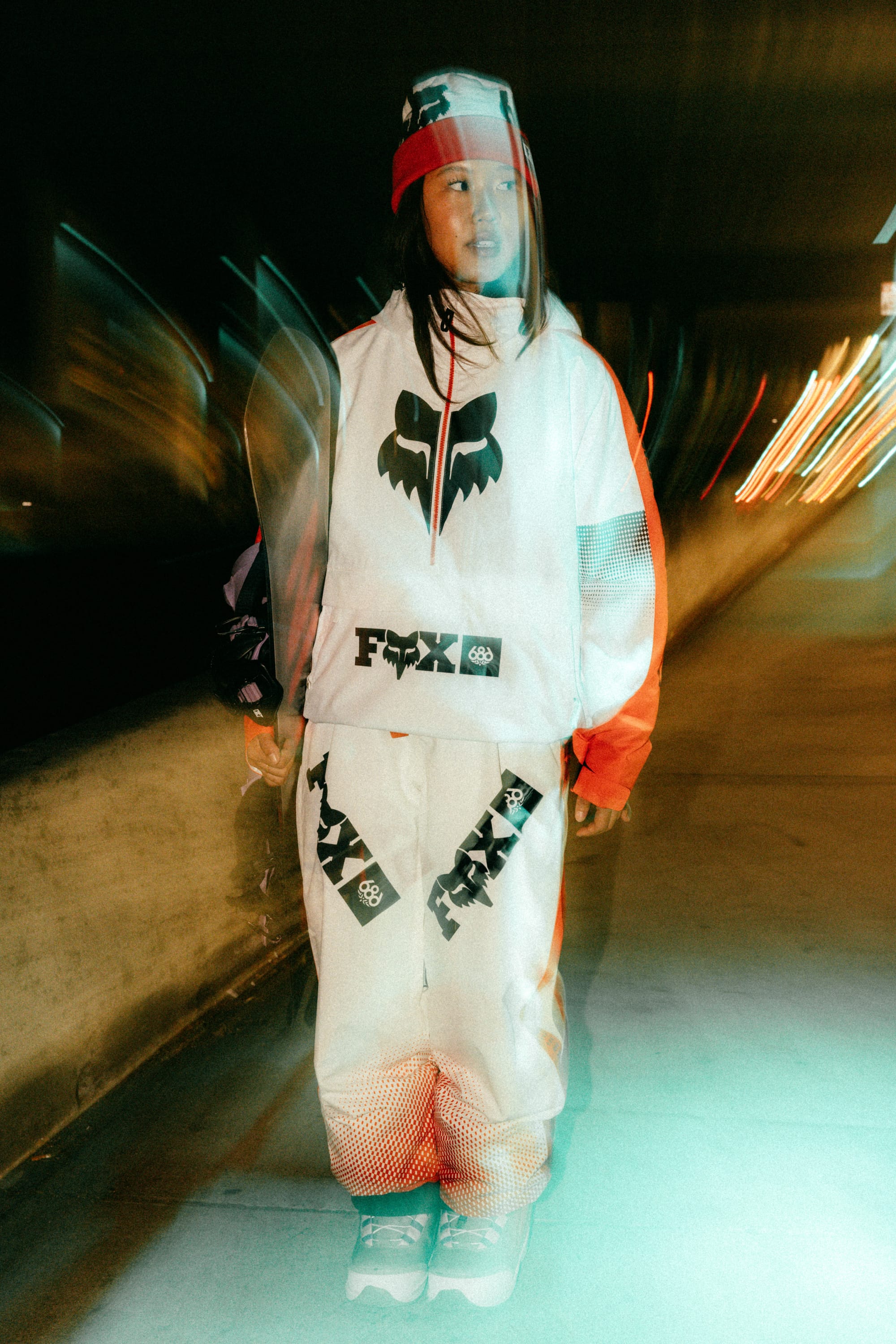 A person stands on a city sidewalk at night wearing 686 Women's Fox Racing Shell Pant, paired with a beanie and sneakers. Light trails and motion blur create a dynamic, urban vibe.