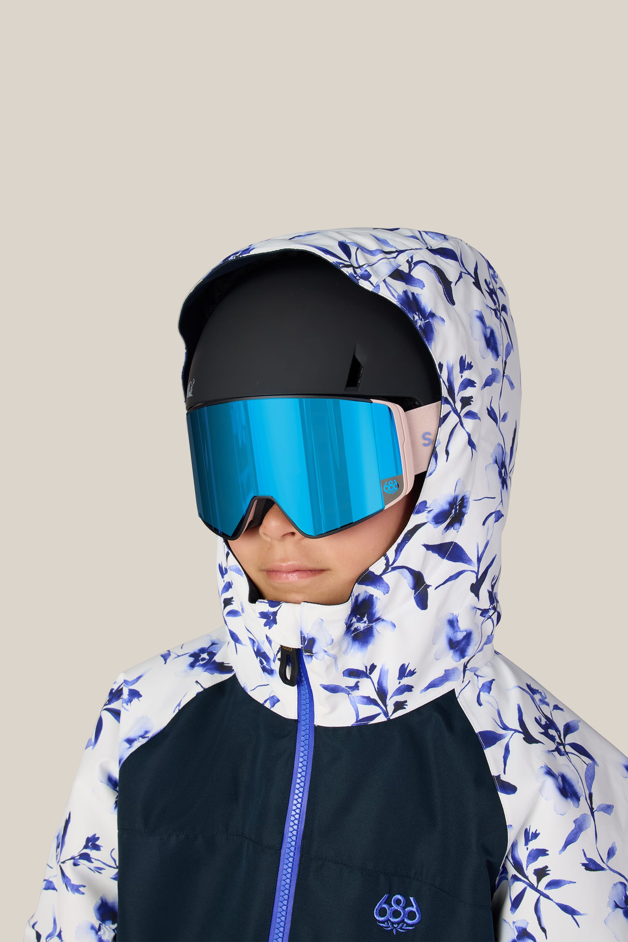 A child wearing the 686 Girls’ Shine Insulated One-Piece stands against a plain beige background, paired with blue reflective ski goggles and a black helmet.
