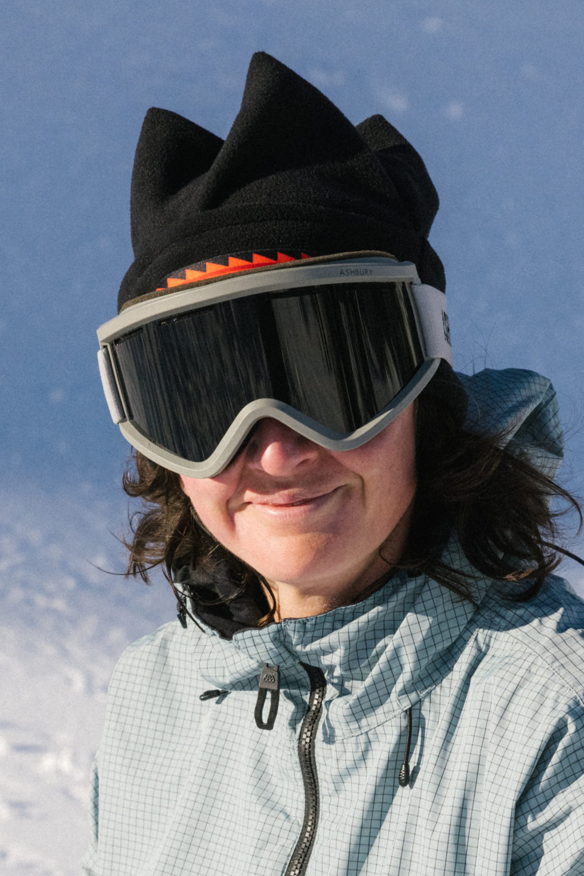A person with long dark hair wears a light checkered jacket, large ski goggles, and the 686 4 Points Fleece Hat by 686, standing outside on a snowy day and smiling at the camera.