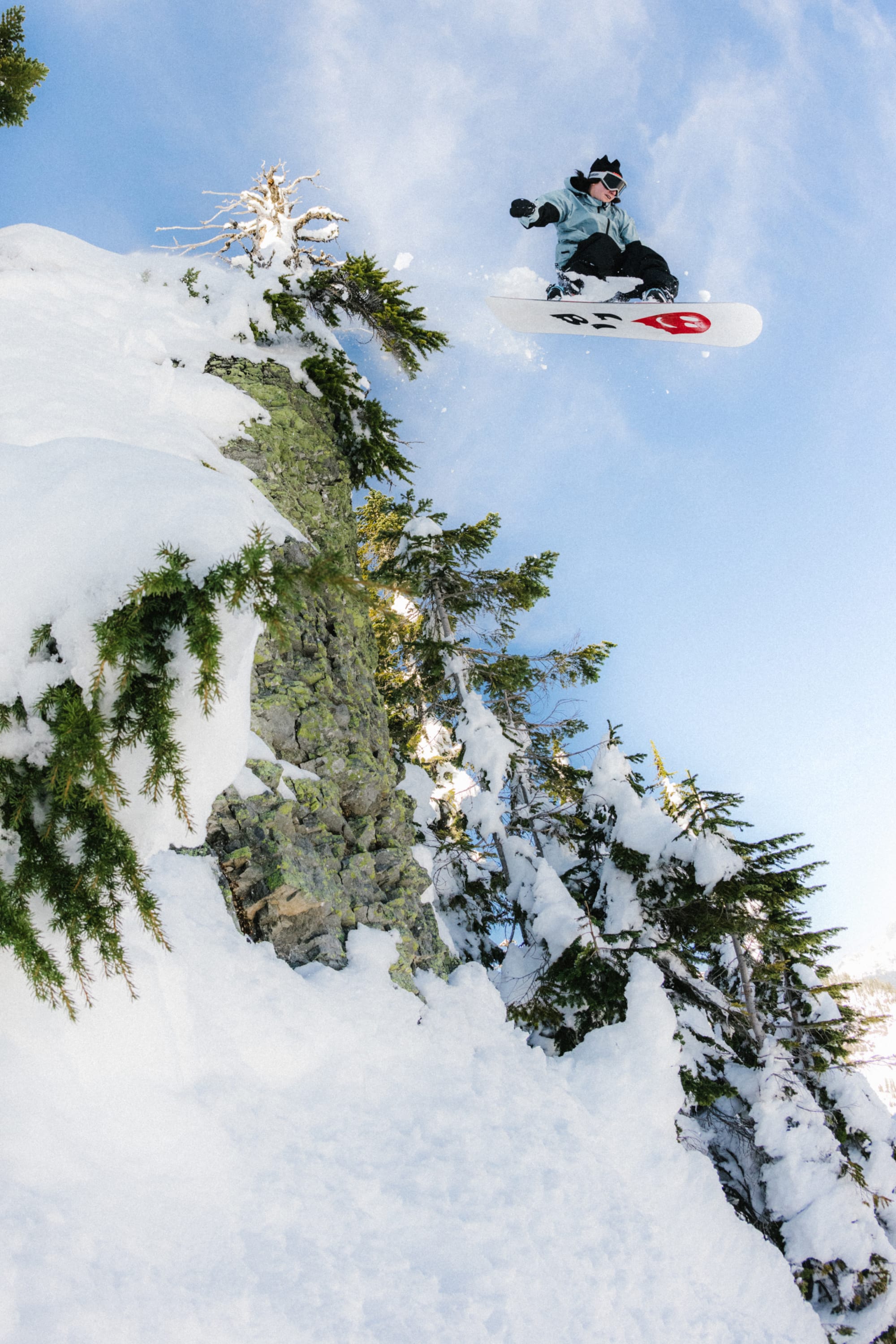 A snowboarder in winter gear and a 686 4 Points Fleece Hat by 686 launches off a snowy, rocky ledge with evergreens, soaring high against a clear blue sky.