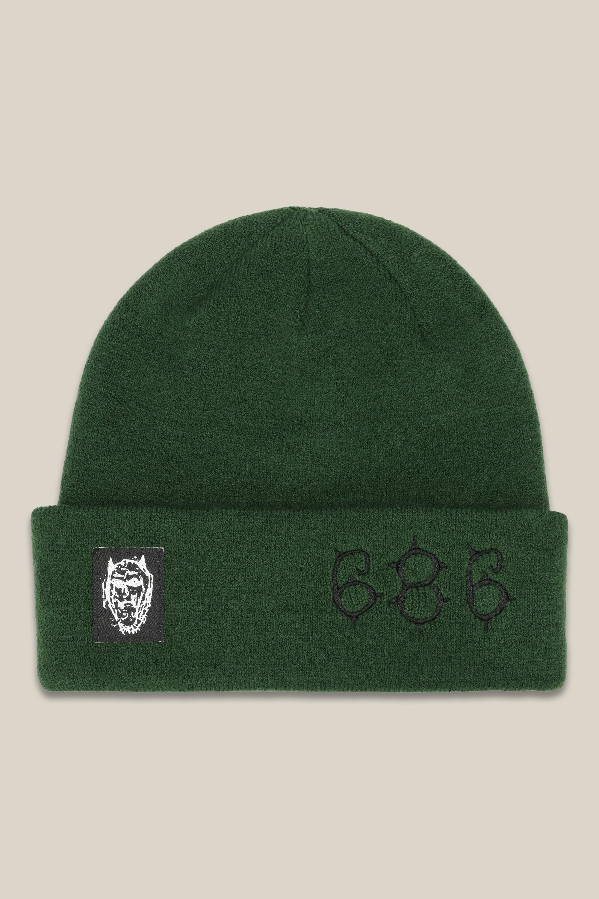 The 686 Cooper Beanie by 686 is a dark green acrylic knit hat with a folded brim, embroidered black 686, and a black-and-white patch featuring an illustrated face on the front.