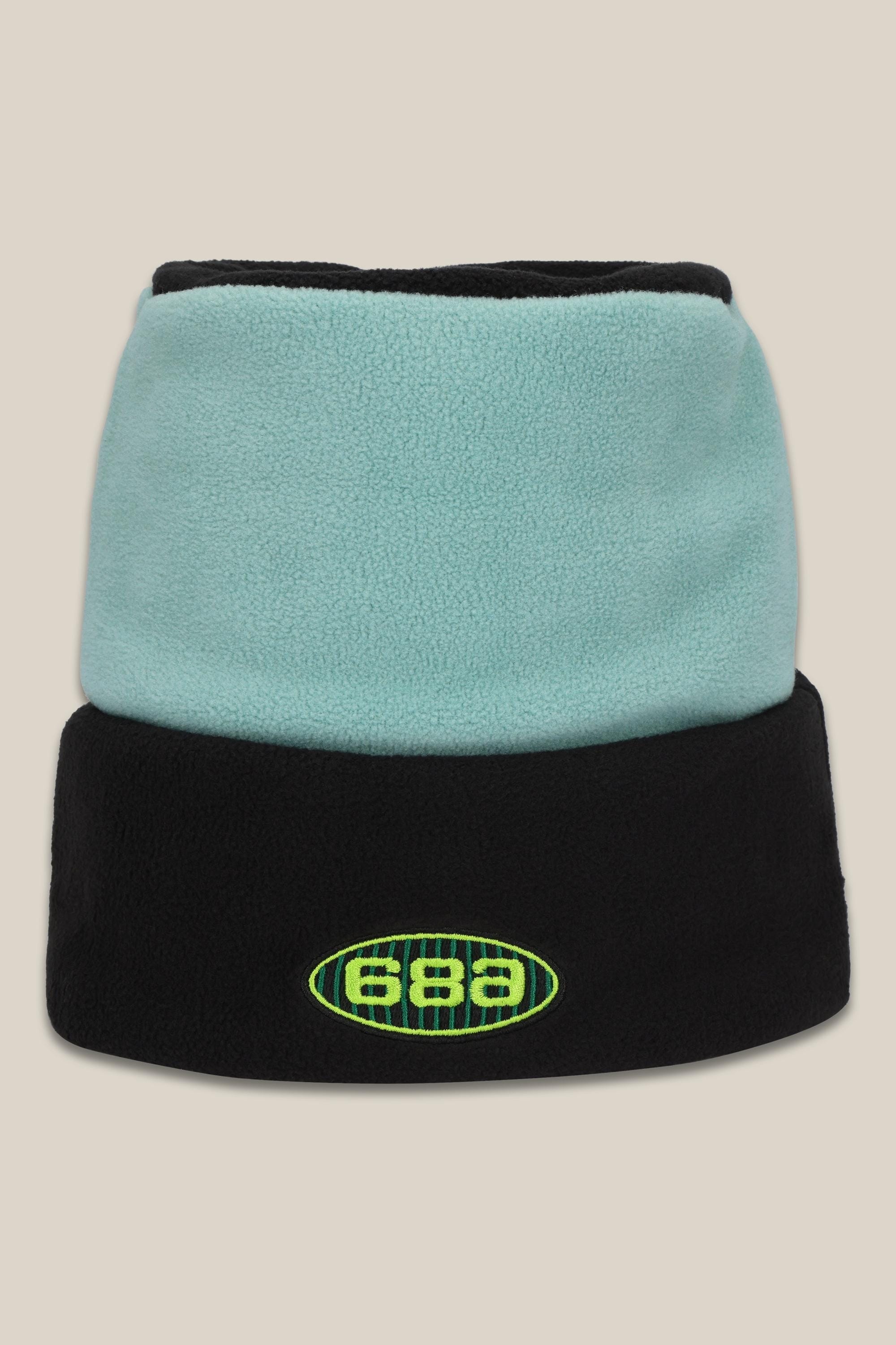 The 686 Flatop Fleece Hat is an acrylic knit beanie with a light blue top, black folded brim, and a green/yellow 686 logo on the front. Set against a plain beige background.
