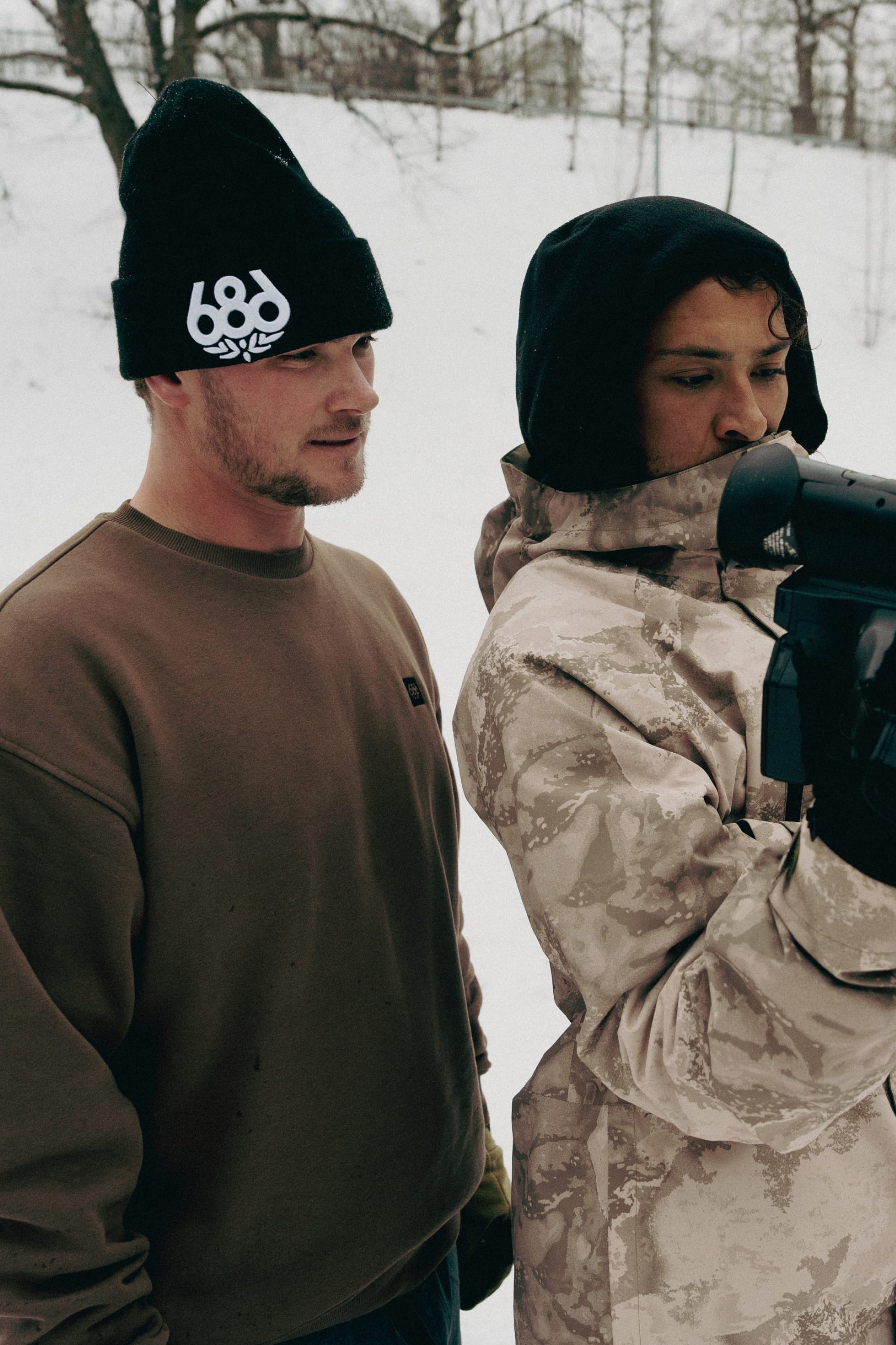 Two people stand outside in snowy surroundings. One wears a brown sweater and the 686 Big Logo Embroidered Beanie by 686, while the other sports a patterned jacket with a black hood and holds a camera or device.