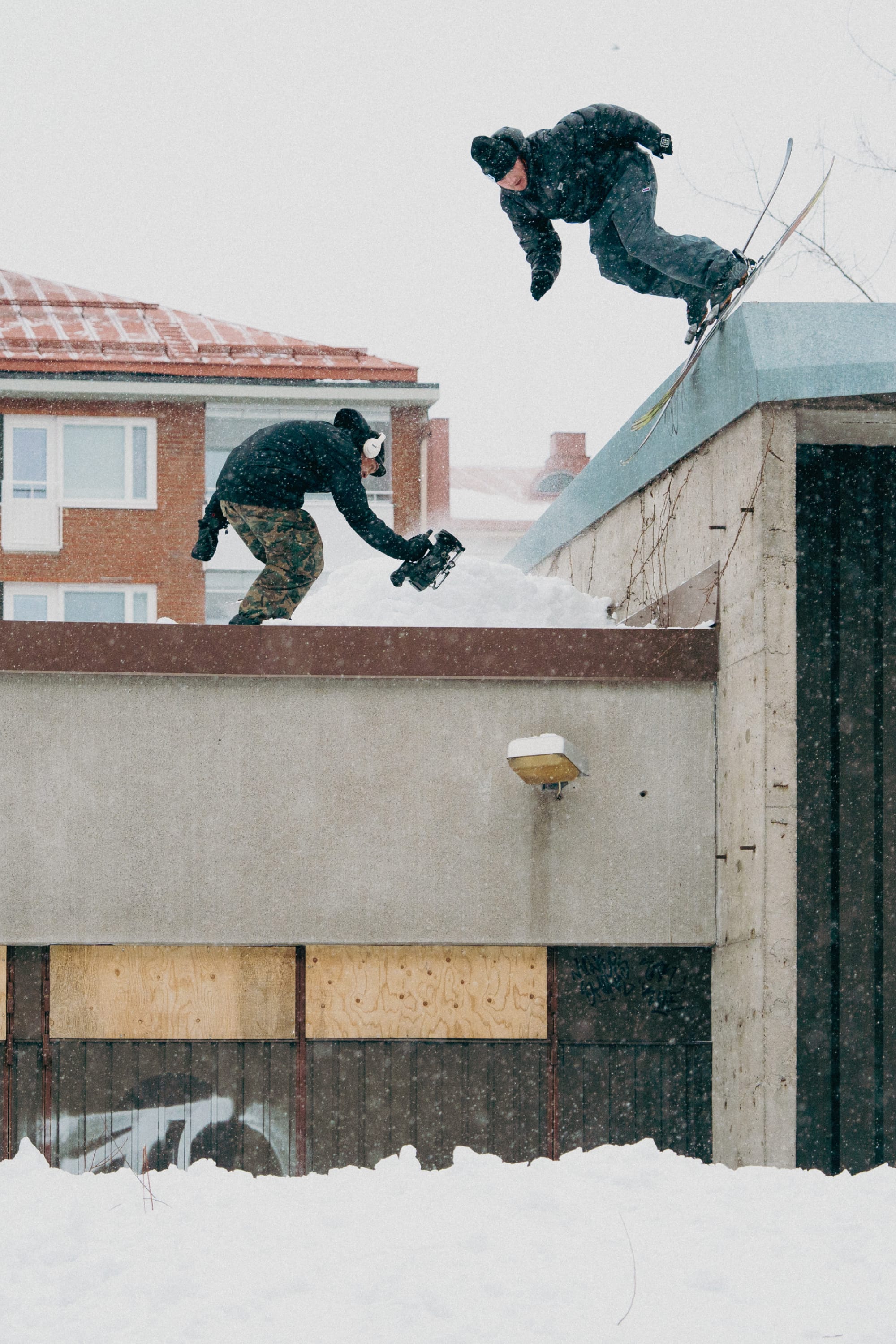 A skier in a 686 Big Logo Embroidered Beanie launches off a snowy rooftop as another films, both dressed for winter amid snow-covered ground and buildings.