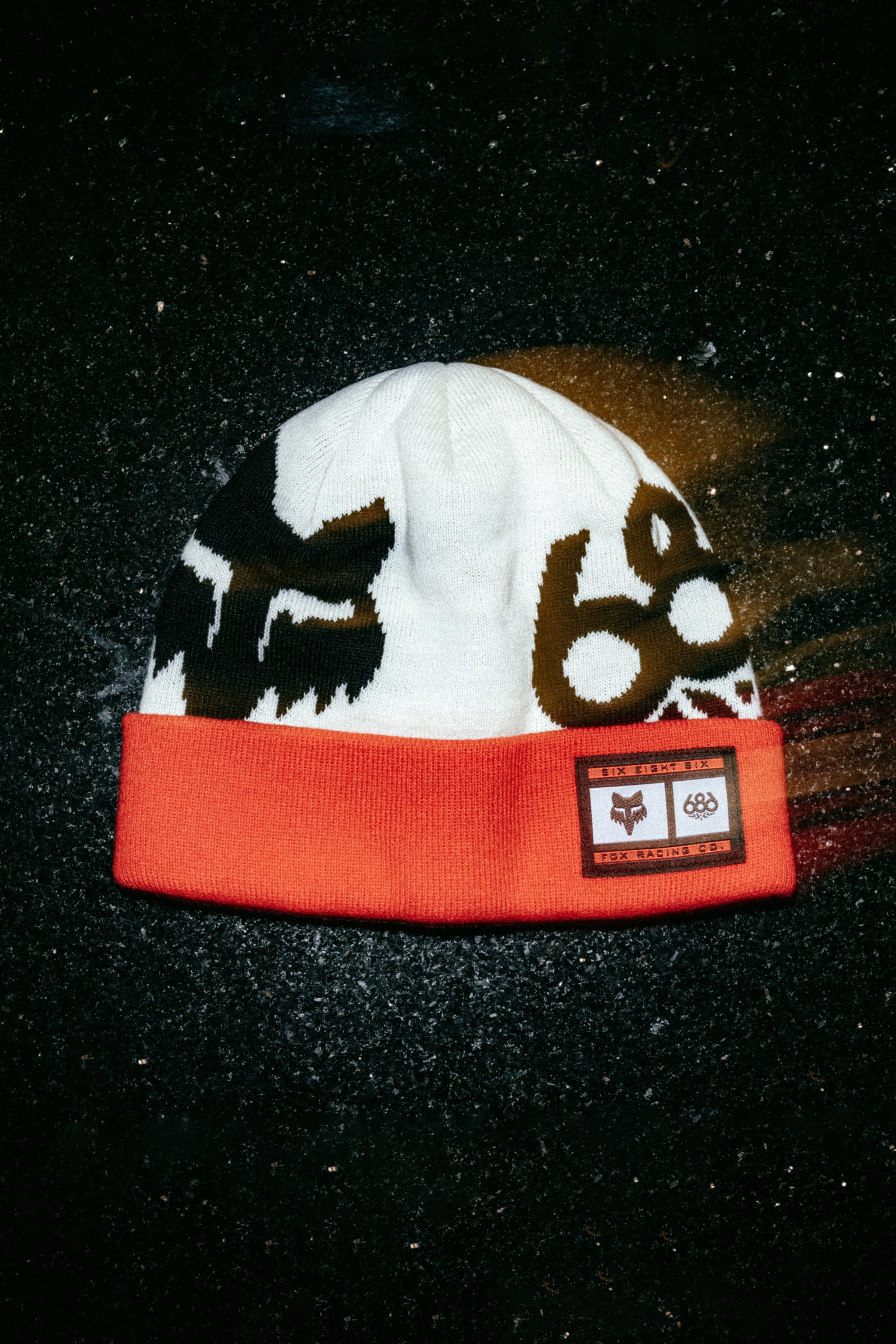 The 686 Fox Racing Cuff Beanie by 686 features a white and red design with black abstract faces and a rectangular front patch, shown against a dark, textured background.