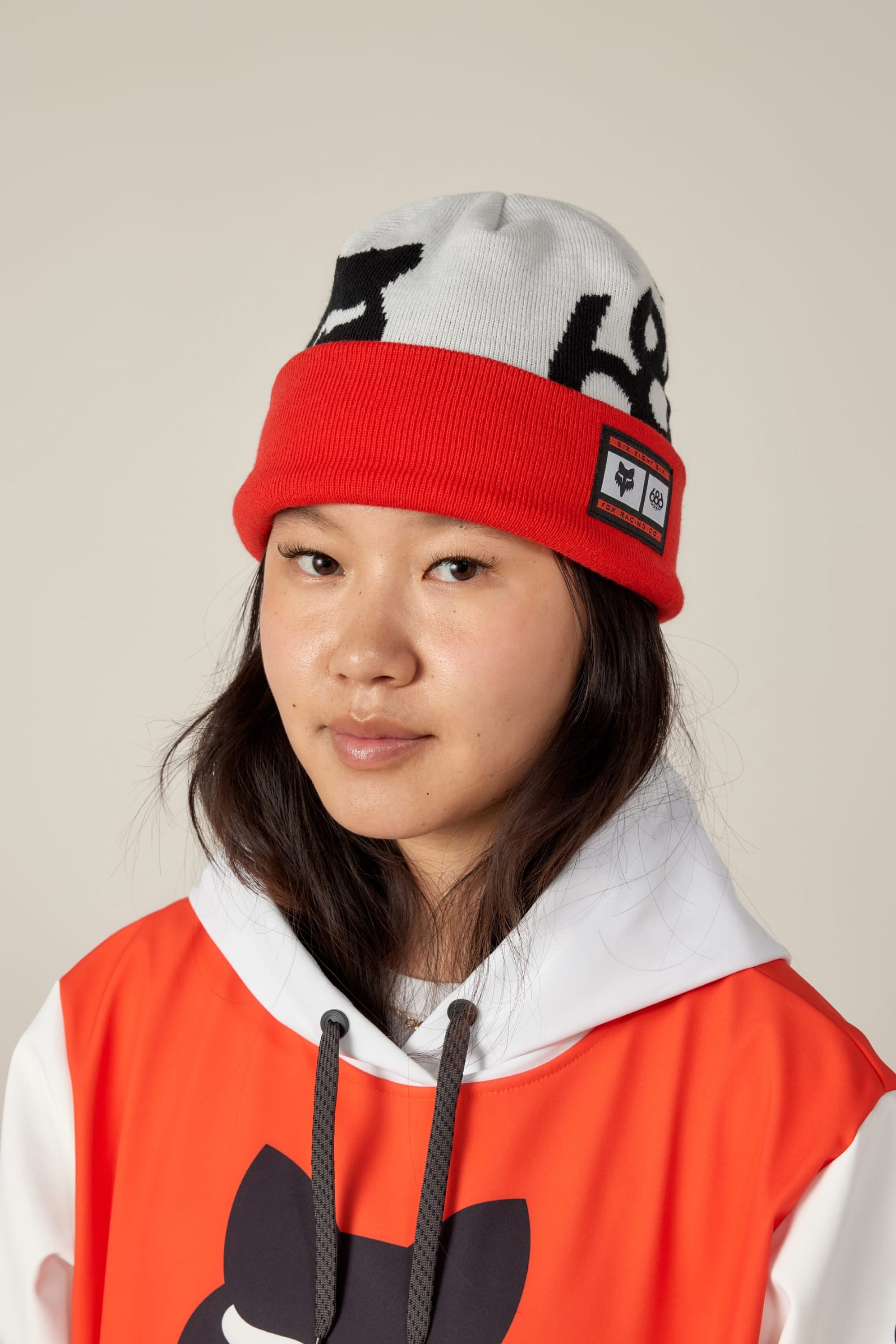 A young woman with long dark hair wears the 686 Fox Racing Cuff Beanie, a white and red hat with a patch and black abstract design by 686, along with an orange and white hoodie with a black graphic on the chest.