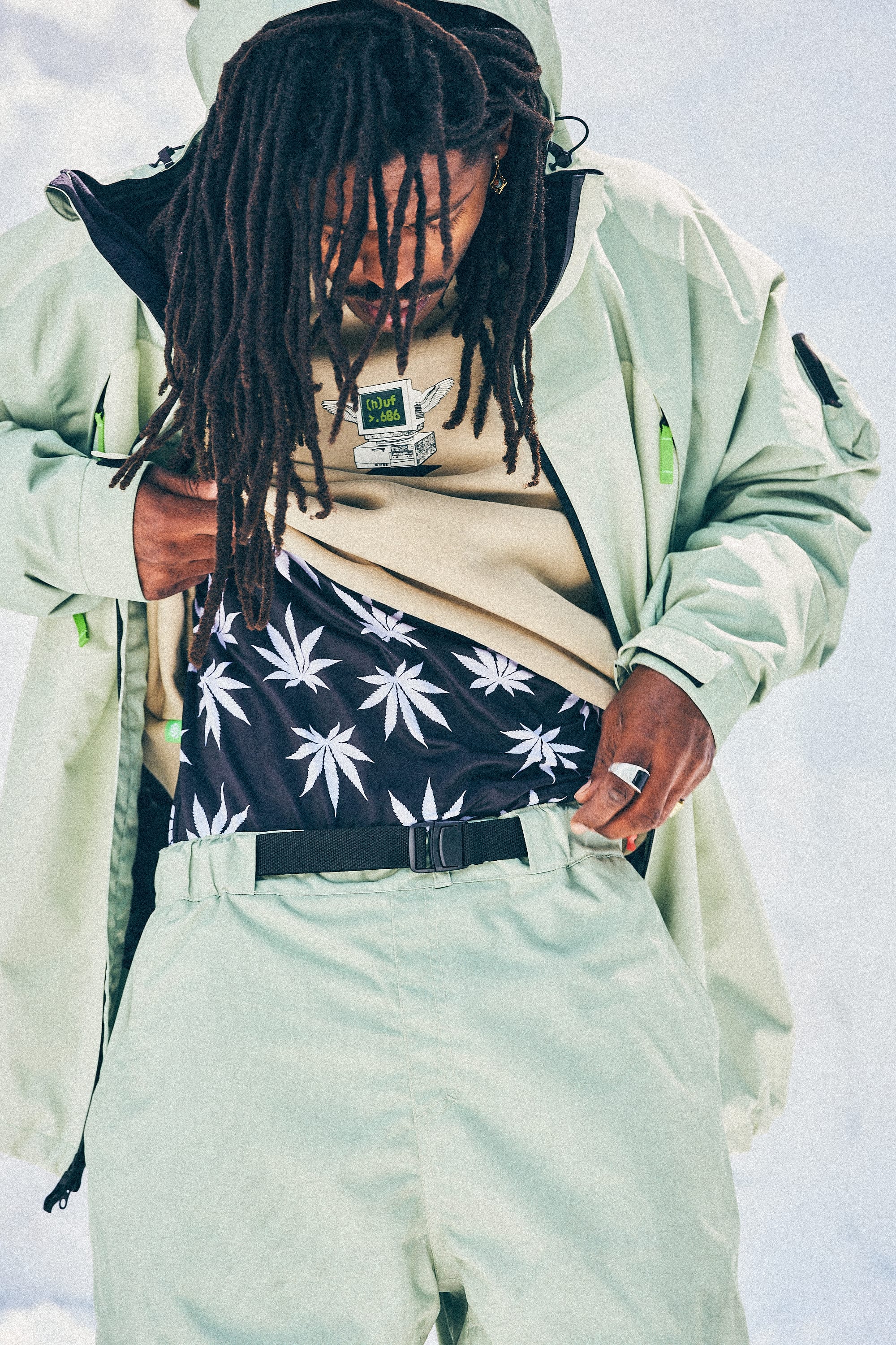 A person with long dreadlocks lifts a beige shirt and mint-green jacket to reveal black shorts with a white cannabis leaf pattern, wearing the 686 Men's HUF Plantlife Base Layer LS Top by 686. The background is bright and sky-like.