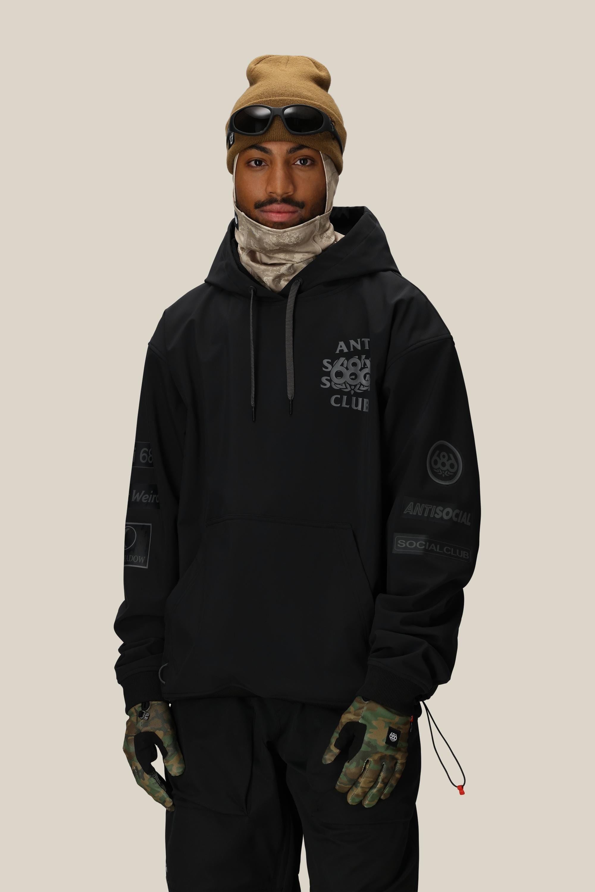 A man wearing a tan beanie with goggles, black pants, camouflage gloves, a beige neck gaiter, and a 686 Men's ASSC F1 Waterproof Team Hoody by 686 stands against a plain light background.