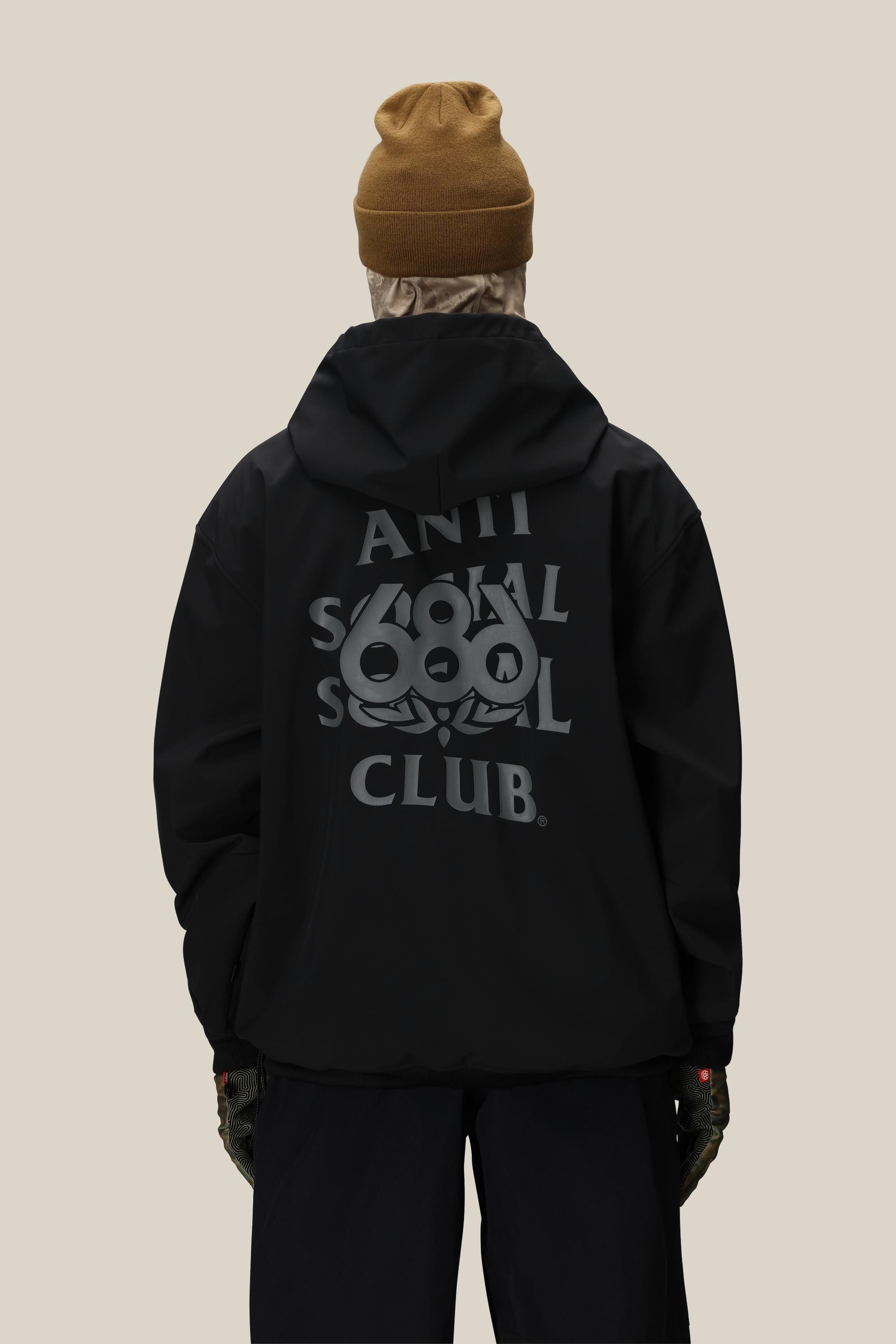 A person wearing a brown beanie, dark pants, gloves, and the 686 Men's ASSC F1 Waterproof Team Hoody by 686, featuring Anti Social Social Club branding and a back graphic, stands facing away against a plain beige background.