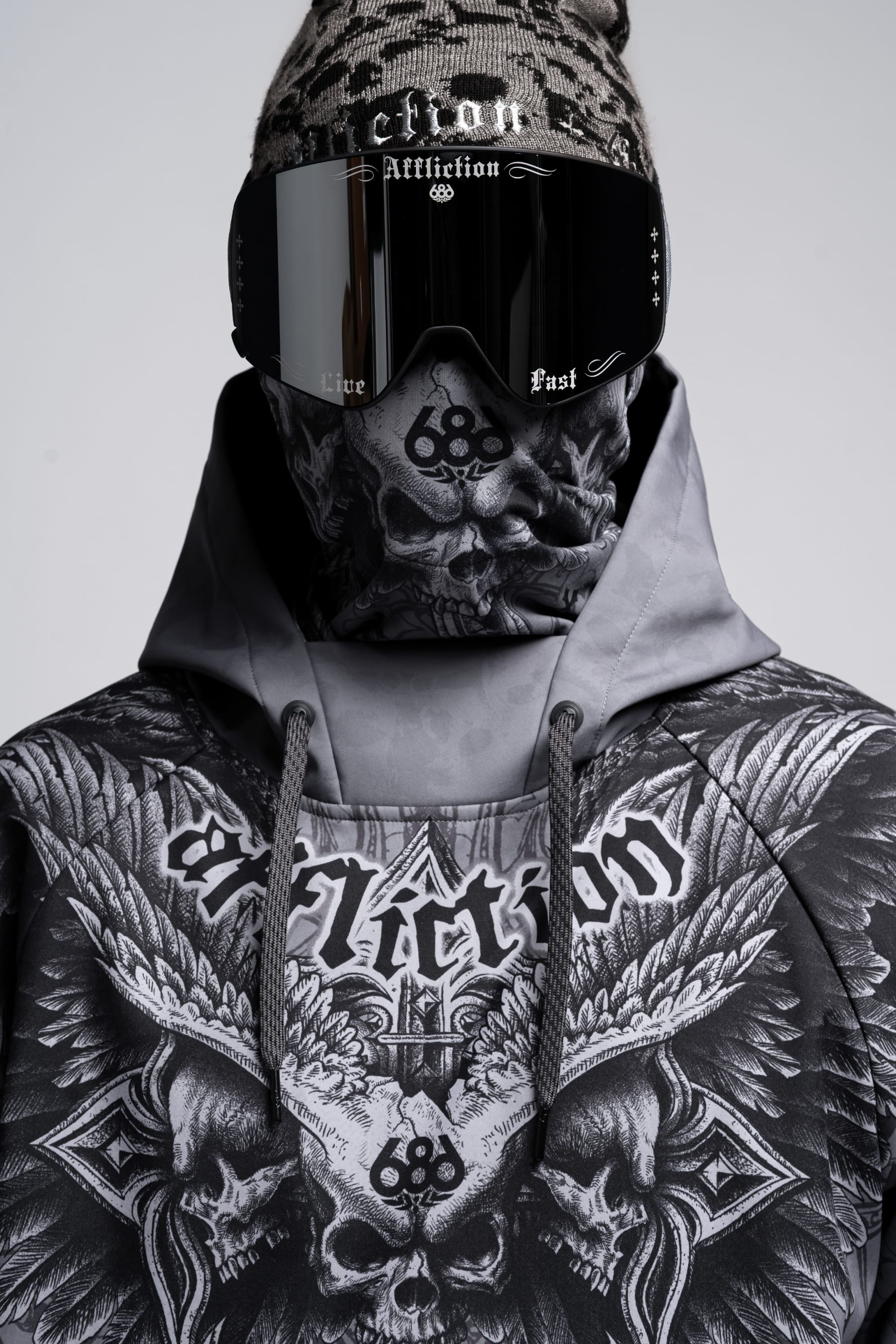 A person in a men's 686 Affliction Waterproof Hoody with intricate skull and wing designs, large black ski goggles, and a beanie. "Affliction" appears boldly on both the 686 hoody and the goggles.