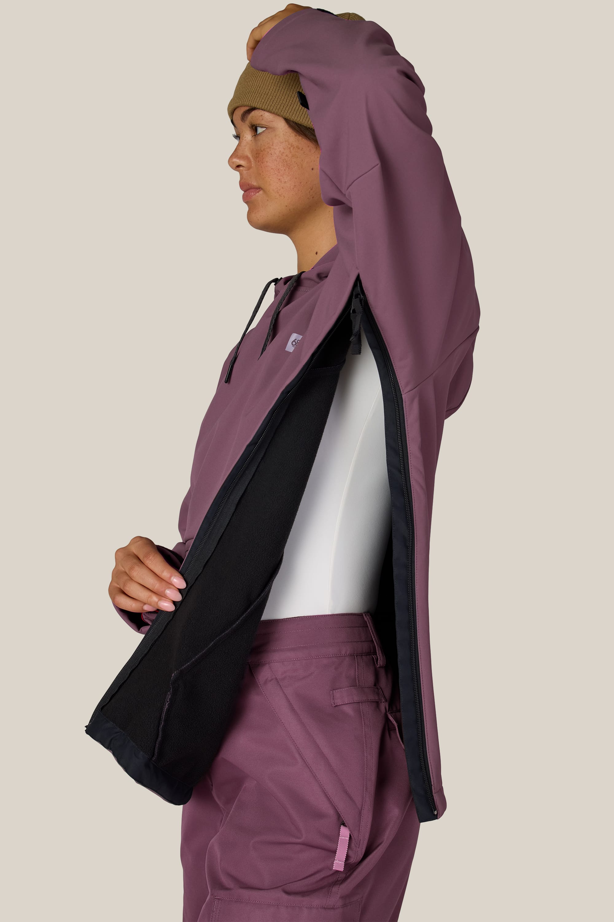 A person in a mauve 686 Women’s Waterproof Hoody and beanie lifts one arm, displaying a large zippered underarm vent lined with black fabric. They stand in profile against a plain, light background.