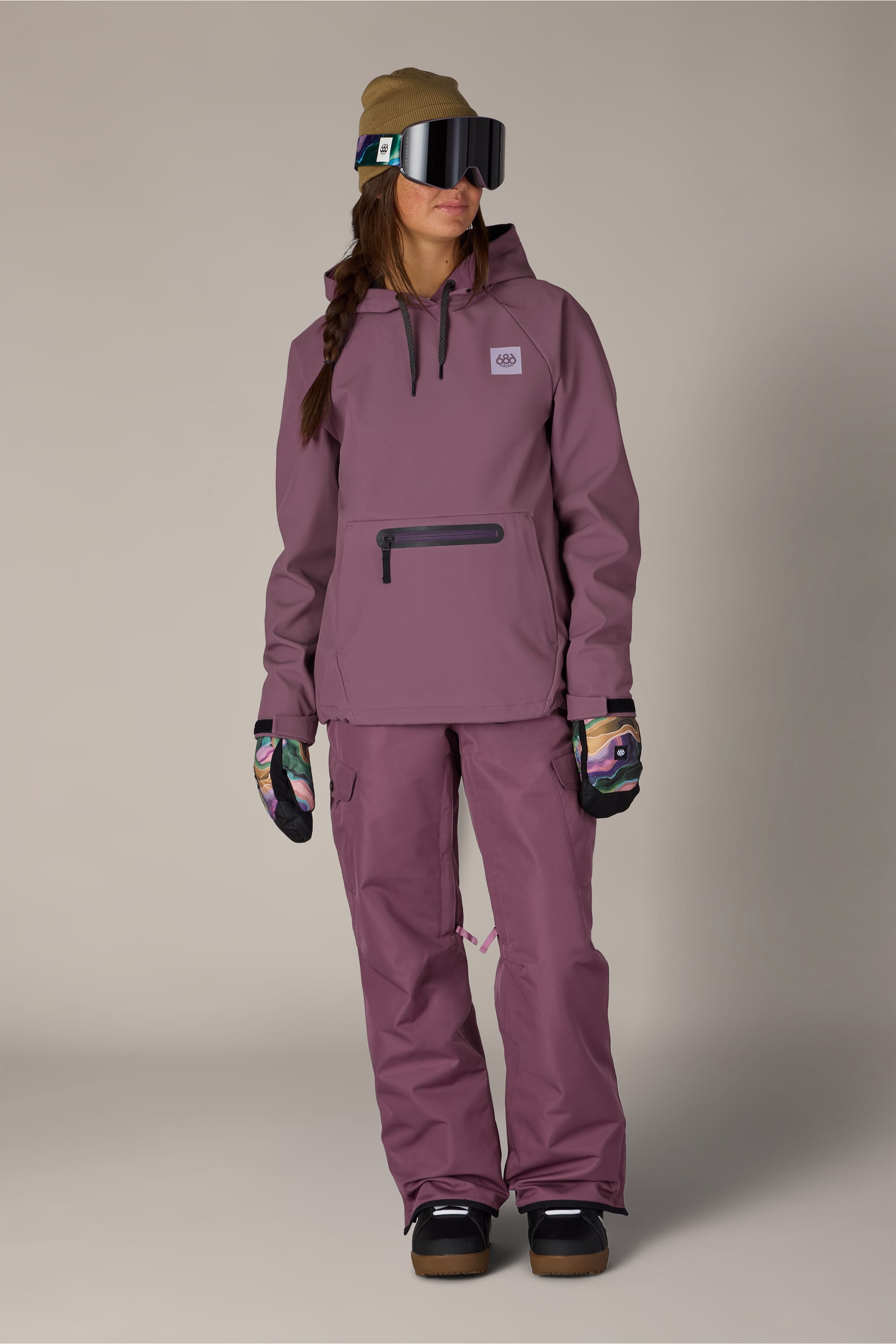 Wearing the 686 Women’s Waterproof Hoody, a person stands facing slightly to the side against a plain background, styled with mauve ski pants, dark gloves, a tan beanie, and large ski goggles.