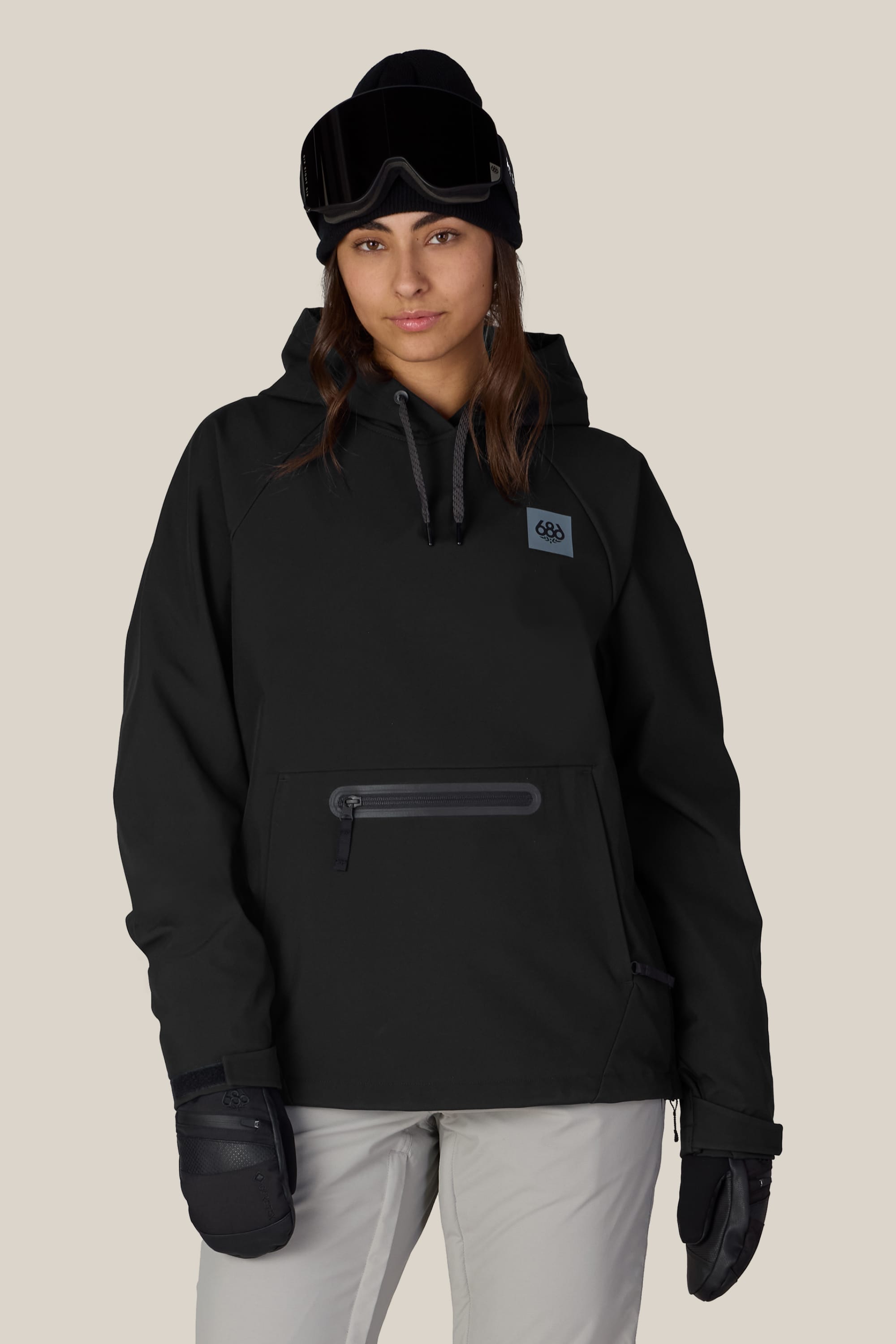 Wearing the 686 Women’s Waterproof Hoody, a person stands facing forward with a neutral expression against a plain background, paired with a beanie, ski goggles, and gloves.