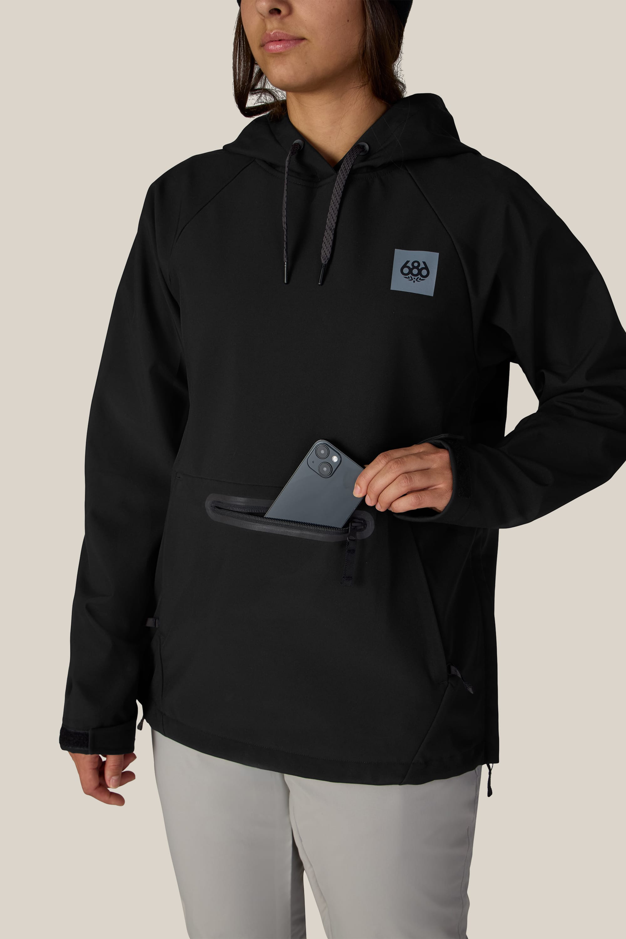 A person wearing the 686 Women’s Waterproof Hoody, featuring a logo on the front, places a smartphone into its front zippered pocket against a plain beige background.
