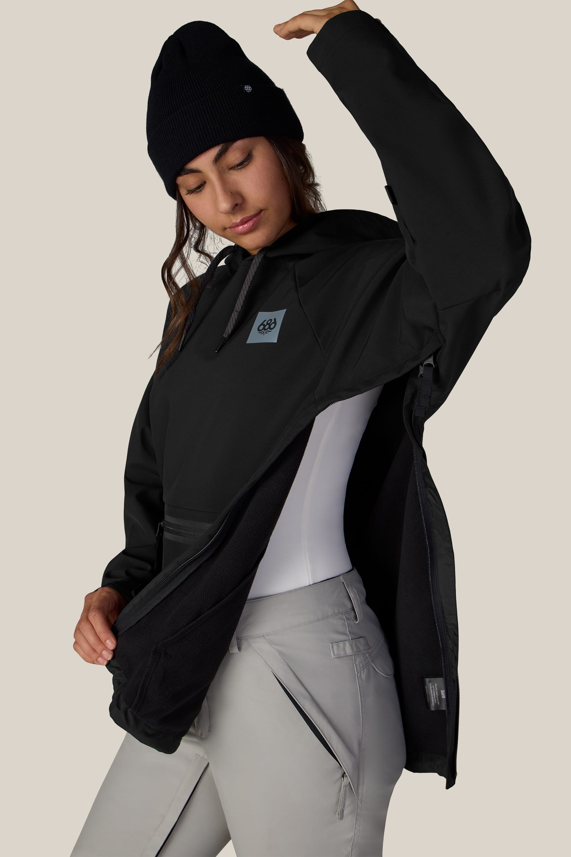 A person wearing the 686 Women’s Waterproof Hoody by 686 in black lifts one arm to reveal the side zipper detail. They layer it over a white shirt and pair it with light gray pants, standing against a plain beige background.