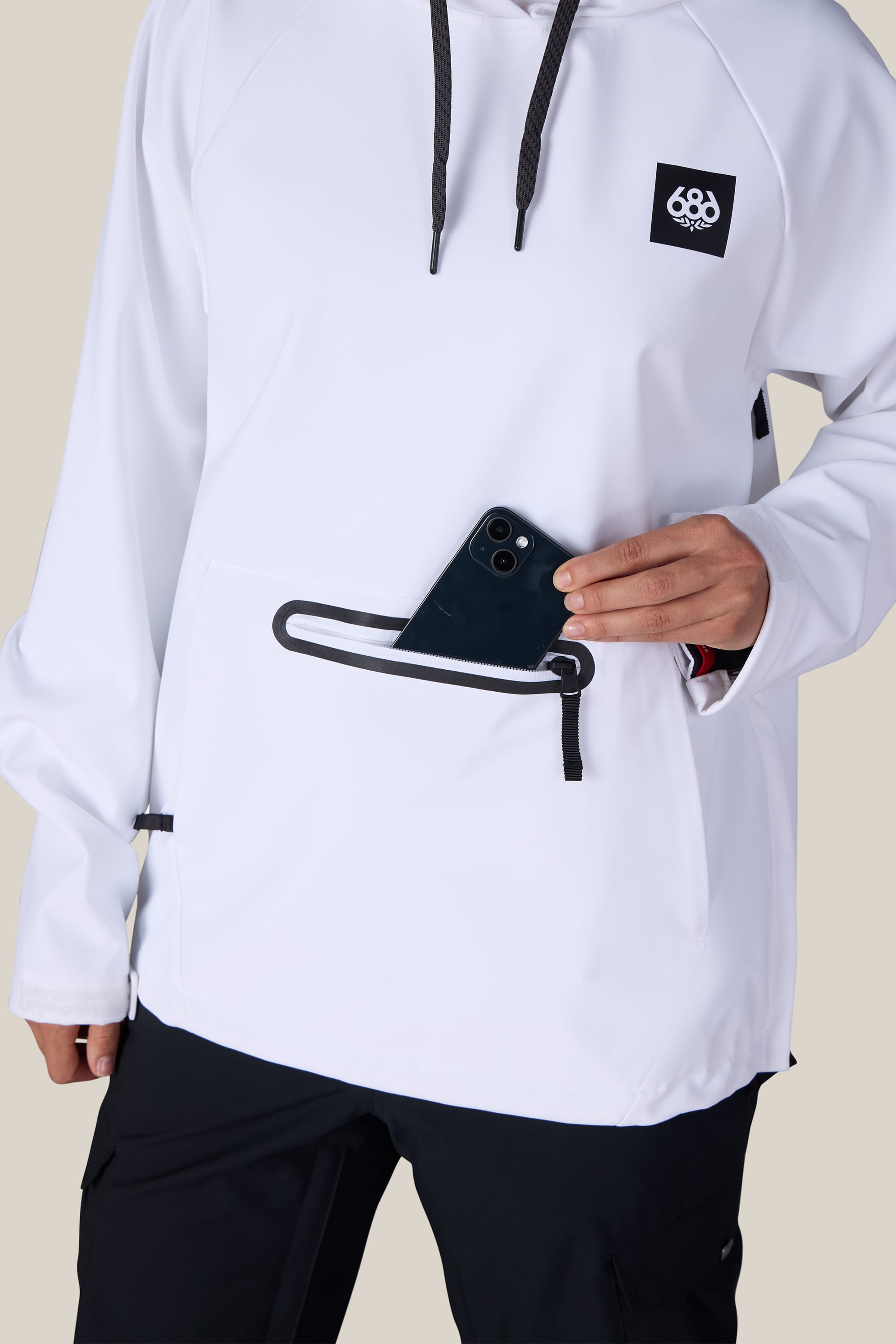 A person wearing the 686 Women’s Waterproof Hoody by 686, featuring black drawstrings and a front zipper pocket, places a black smartphone inside the pocket. Only their torso and one hand are visible.