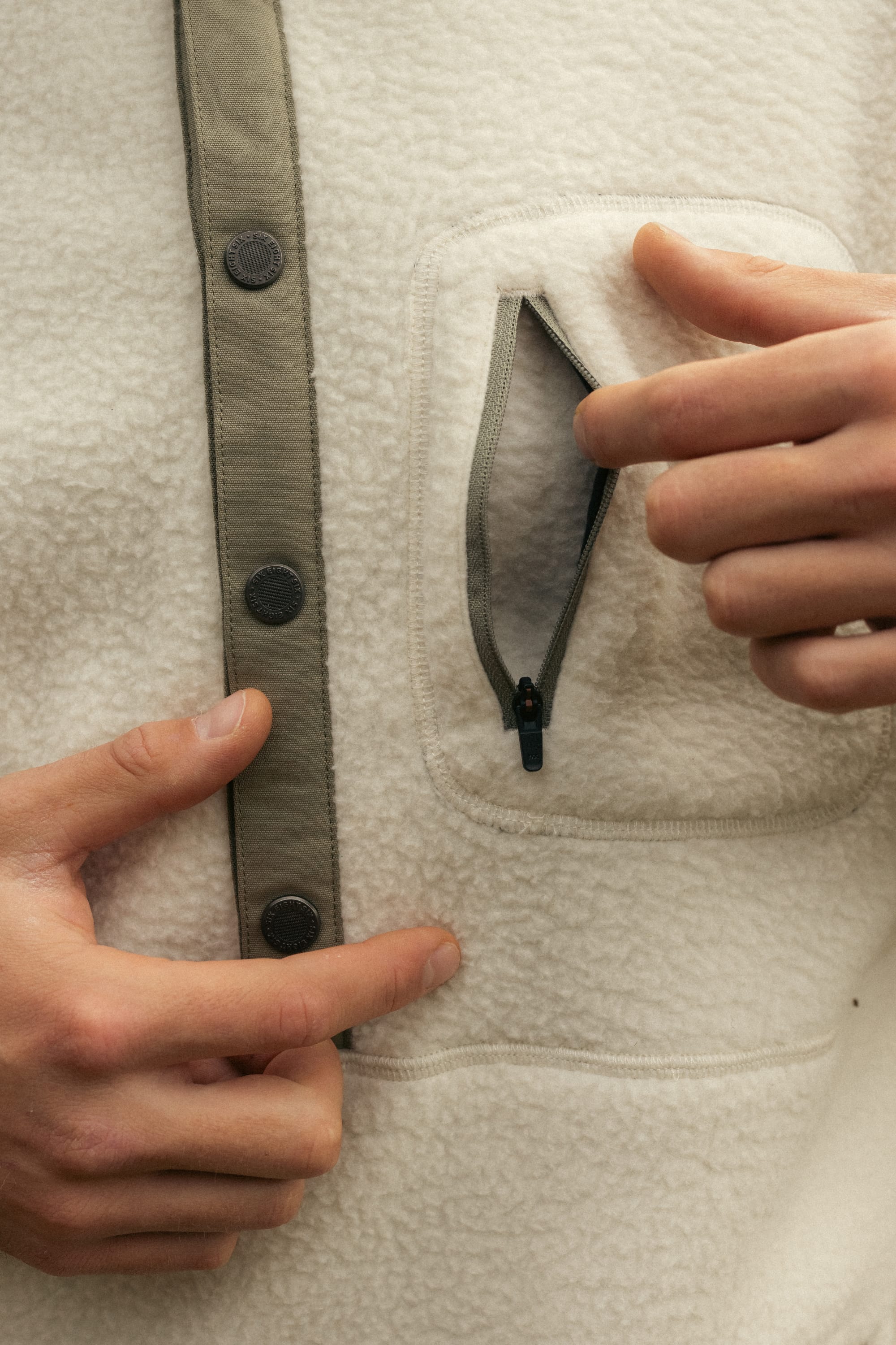 A man opens a small zippered chest pocket on the cream-colored 686 Mens Tioga Fleece Pullover, featuring dark buttons and trim.