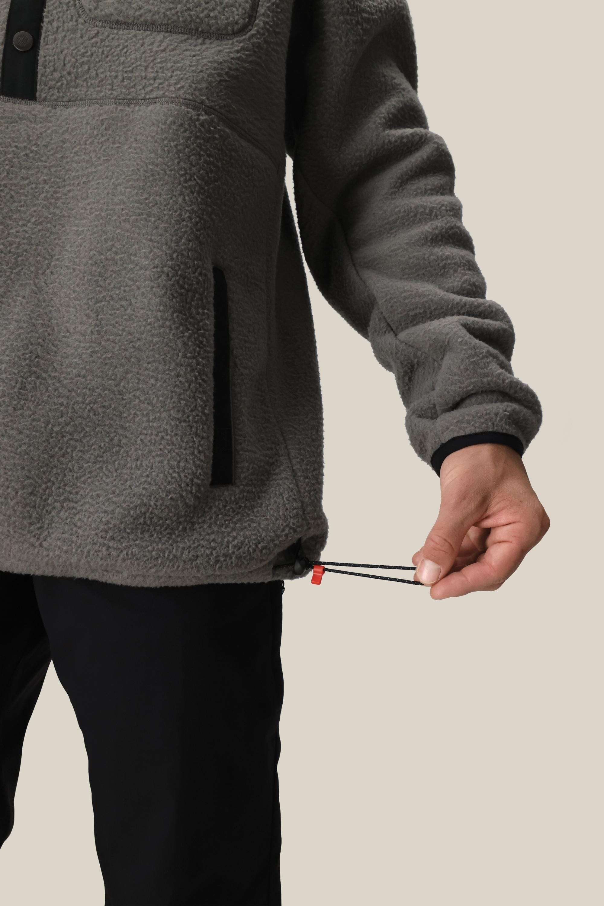 A person in a gray 686 Mens Tioga Fleece Pullover adjusts the hem drawstring with their right hand. Only their lower torso and arm are visible against a plain beige background.