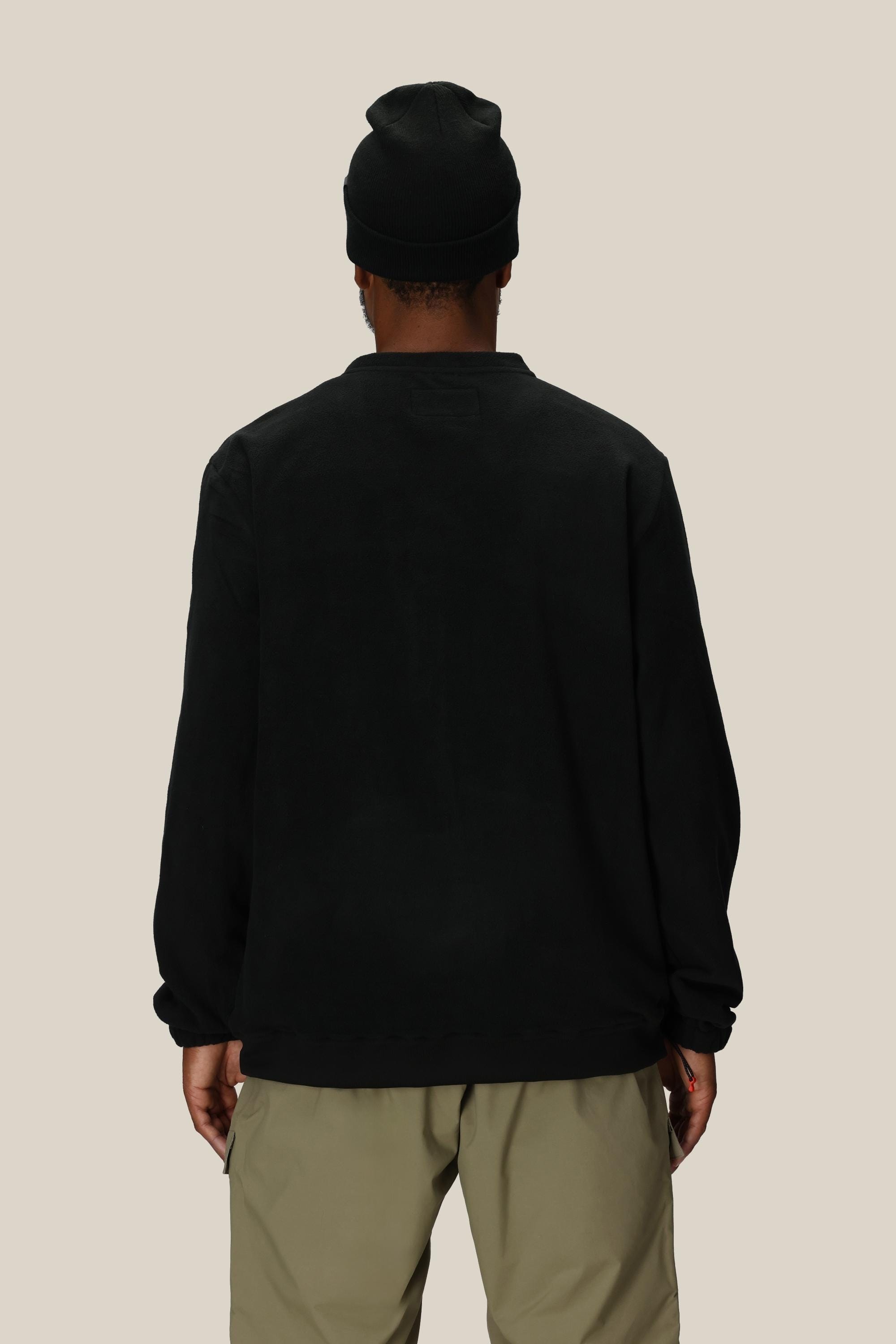 A person wearing a black beanie, the 686 Mens Mystery Ranch Qi Fleece, and light khaki pants stands with their back to the camera against a plain beige background.