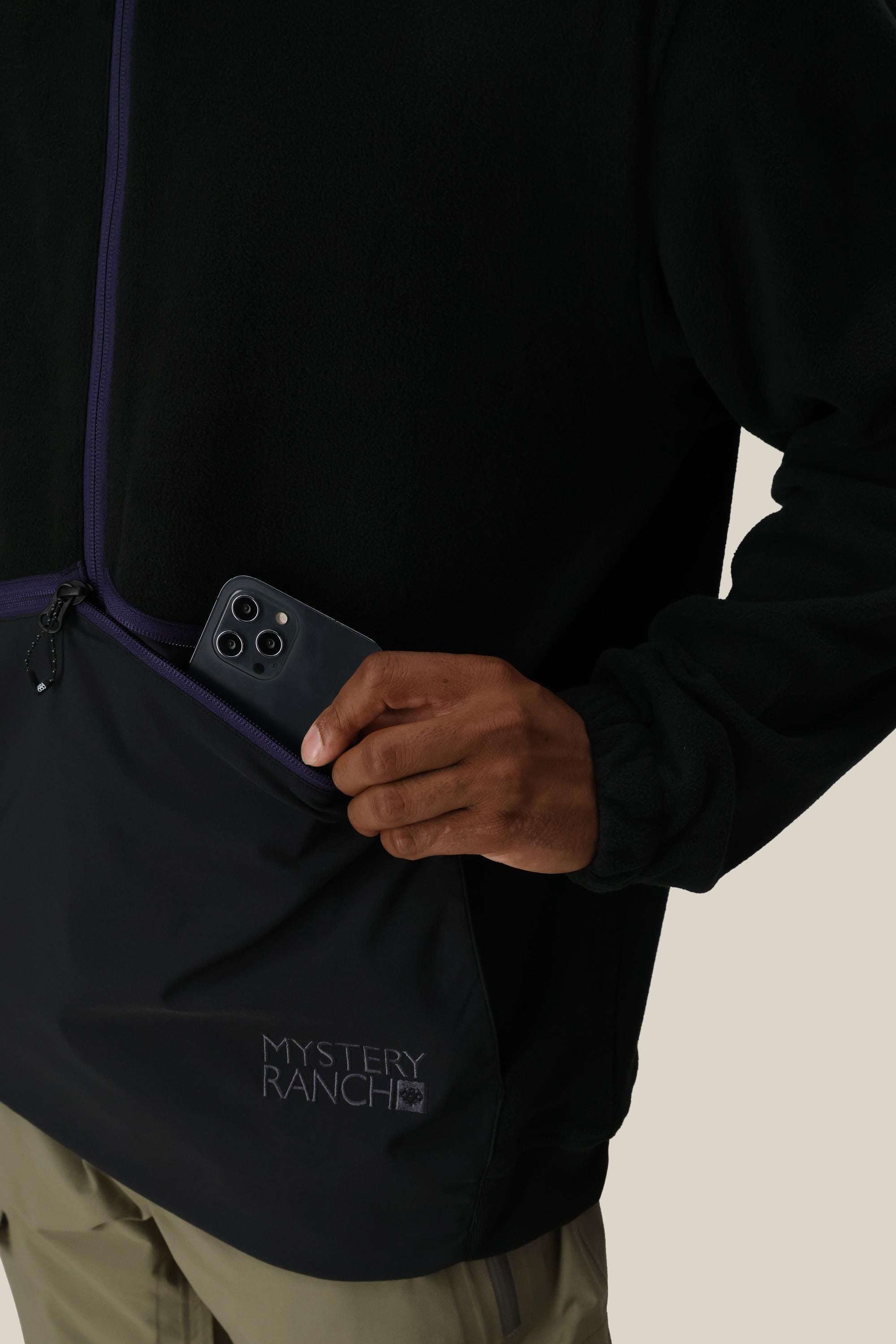 A person wearing the 686 Mens Mystery Ranch Qi Fleece in black is placing a black smartphone into its zippered front pocket. The fleece features Mystery Ranch branding near the pocket and is paired with light brown pants.