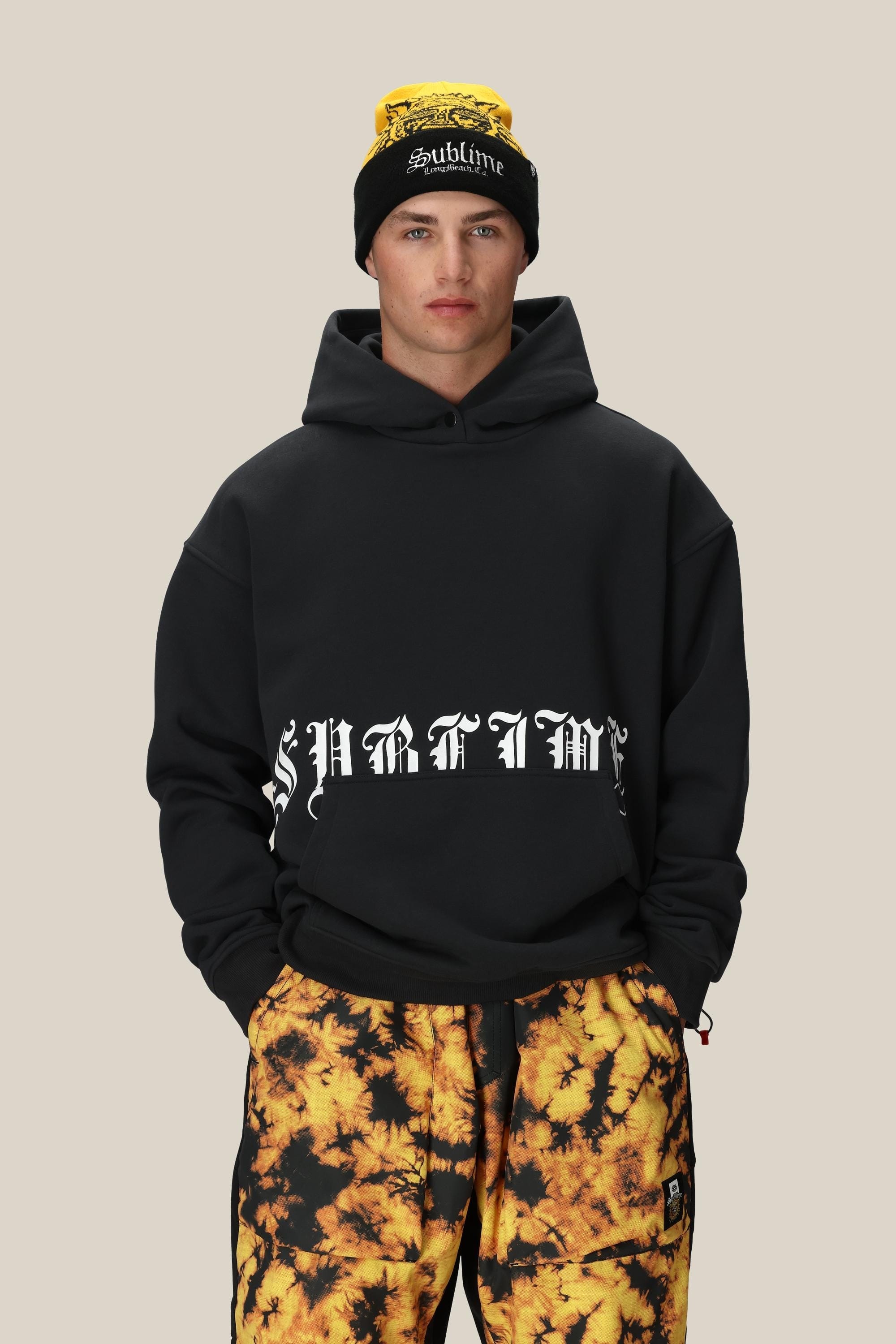 A person wears the 686 Men's Sublime Premium Heavyweight Pullover Hoody in black, paired with a black beanie with yellow "Sublime" text and yellow-black tie-dye pants, standing against a plain beige background.