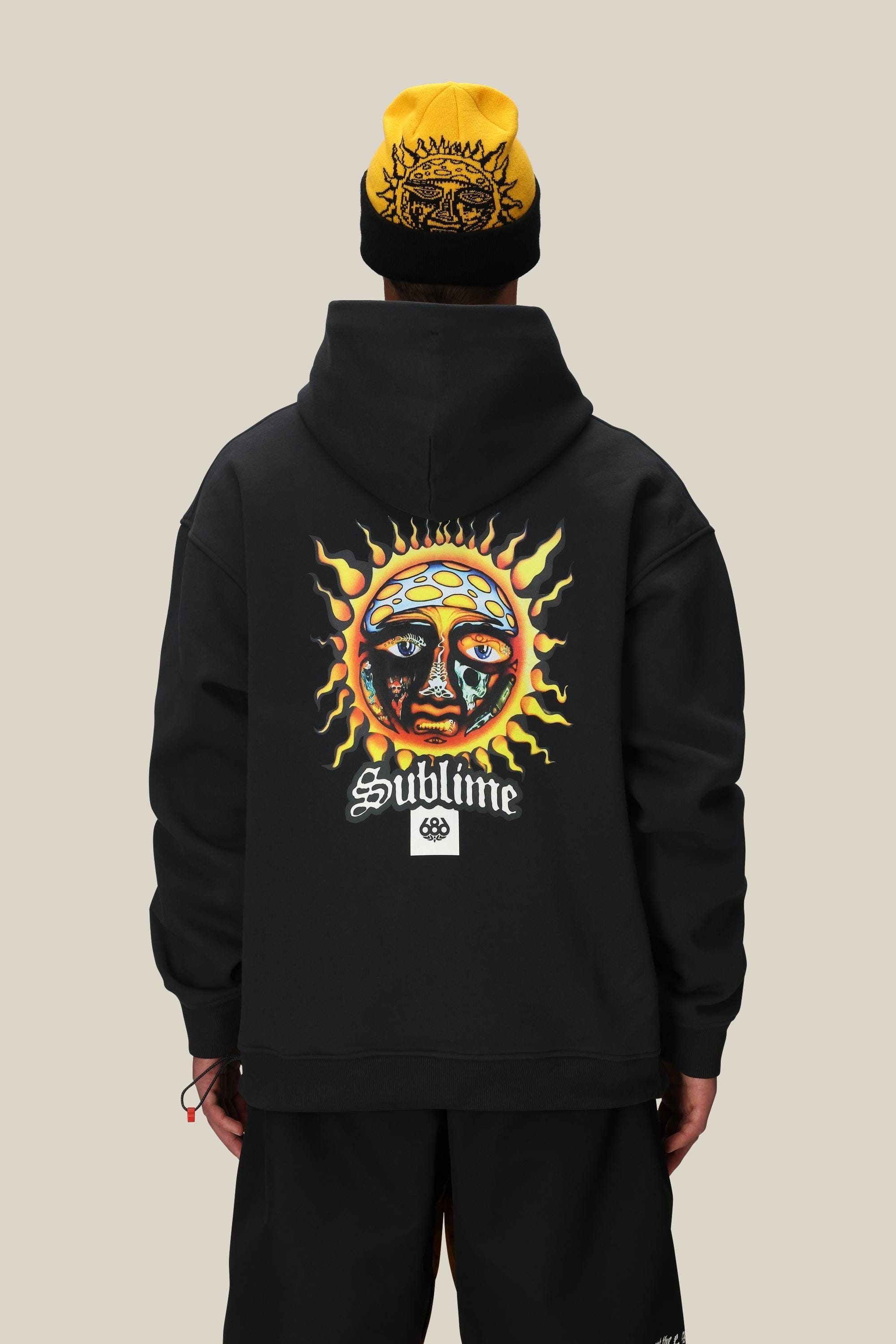 A person wears the 686 Men's Sublime Premium Heavyweight Pullover Hoody featuring a colorful sun and face graphic with "Sublime" on the back, paired with a black and yellow beanie, standing against a plain beige background.