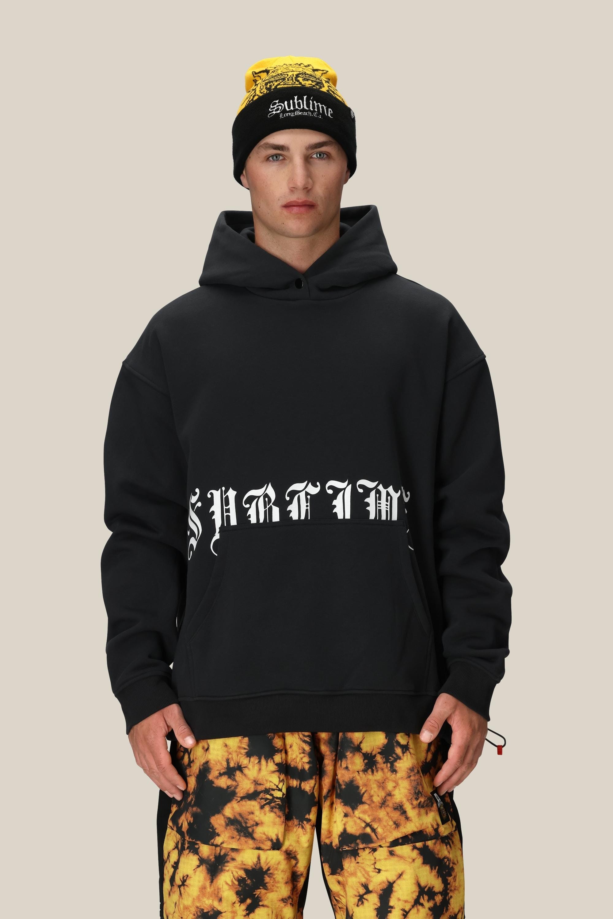 A person in the 686 Men's Sublime Premium Heavyweight Pullover Hoody and a black "Sublime" beanie stands against a beige background, paired with yellow-black tie-dye pants.