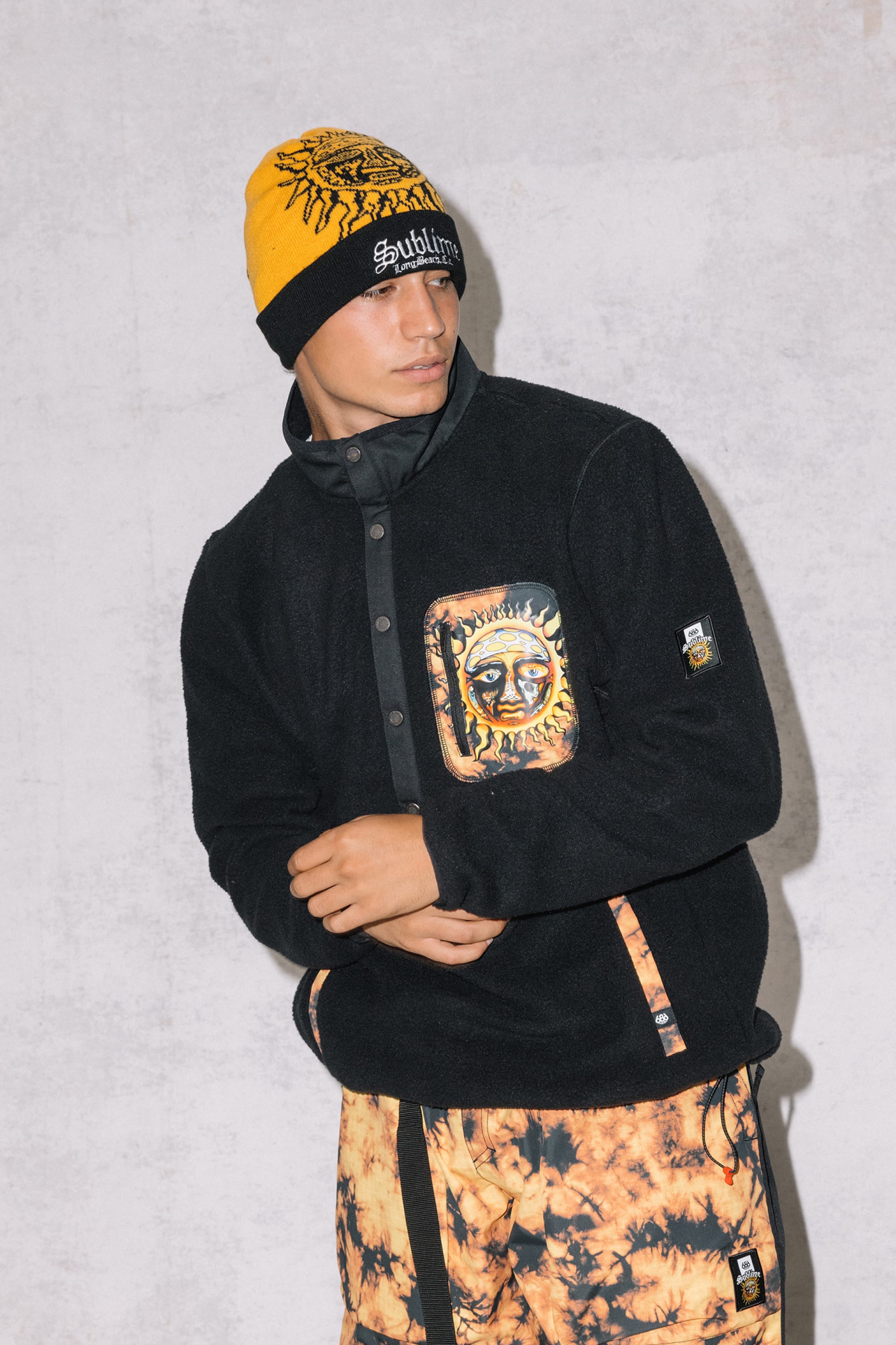 A person wearing the 686 Mens Sublime Tioga Fleece Pullover stands against a light gray textured background, paired with a yellow and black knit beanie and tie-dye yellow and black pants.