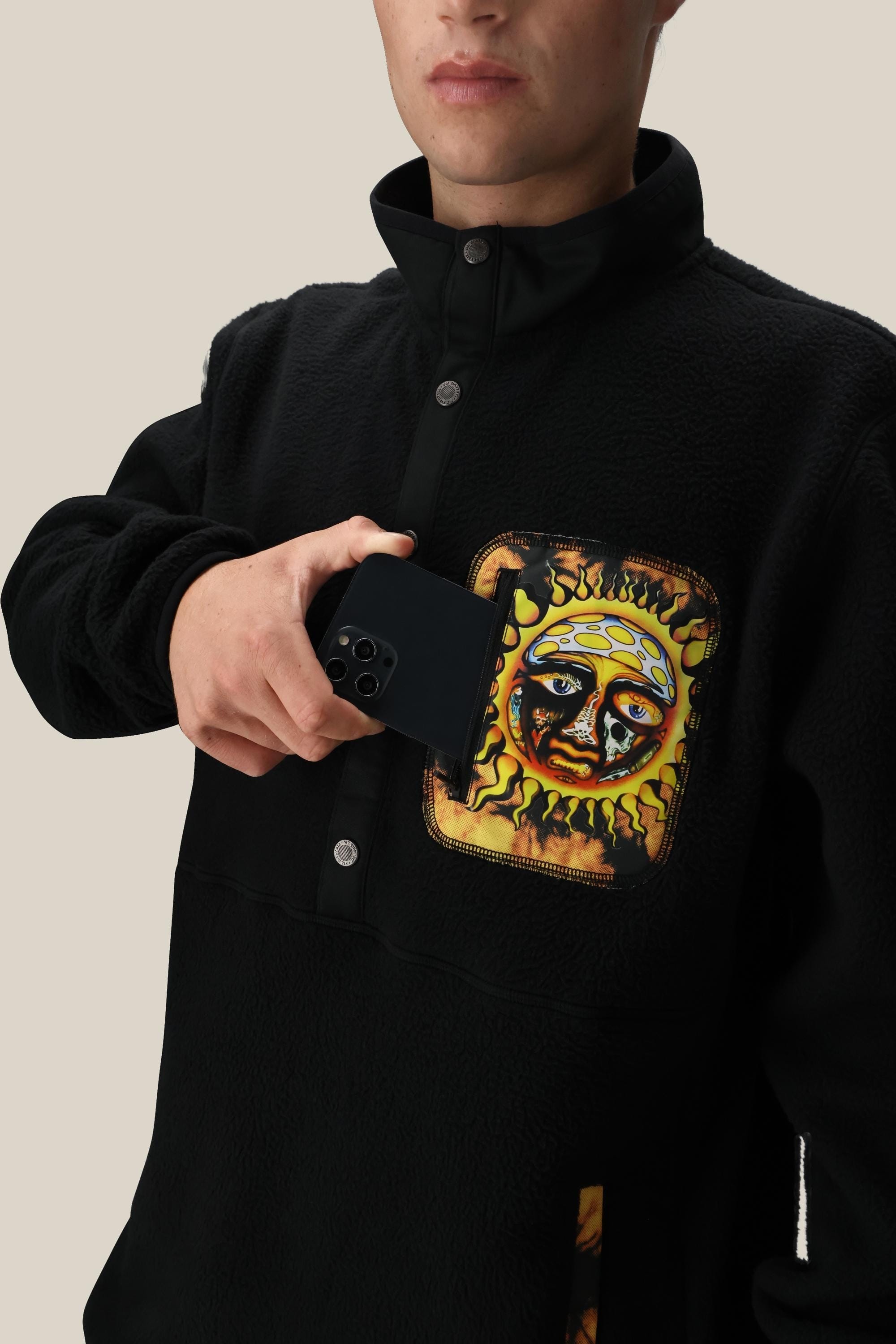 A person wears the 686 Mens Sublime Tioga Fleece Pullover by 686, featuring a colorful sun face design on the chest pocket, and holds a black smartphone partially inside the pocket against a plain beige background.