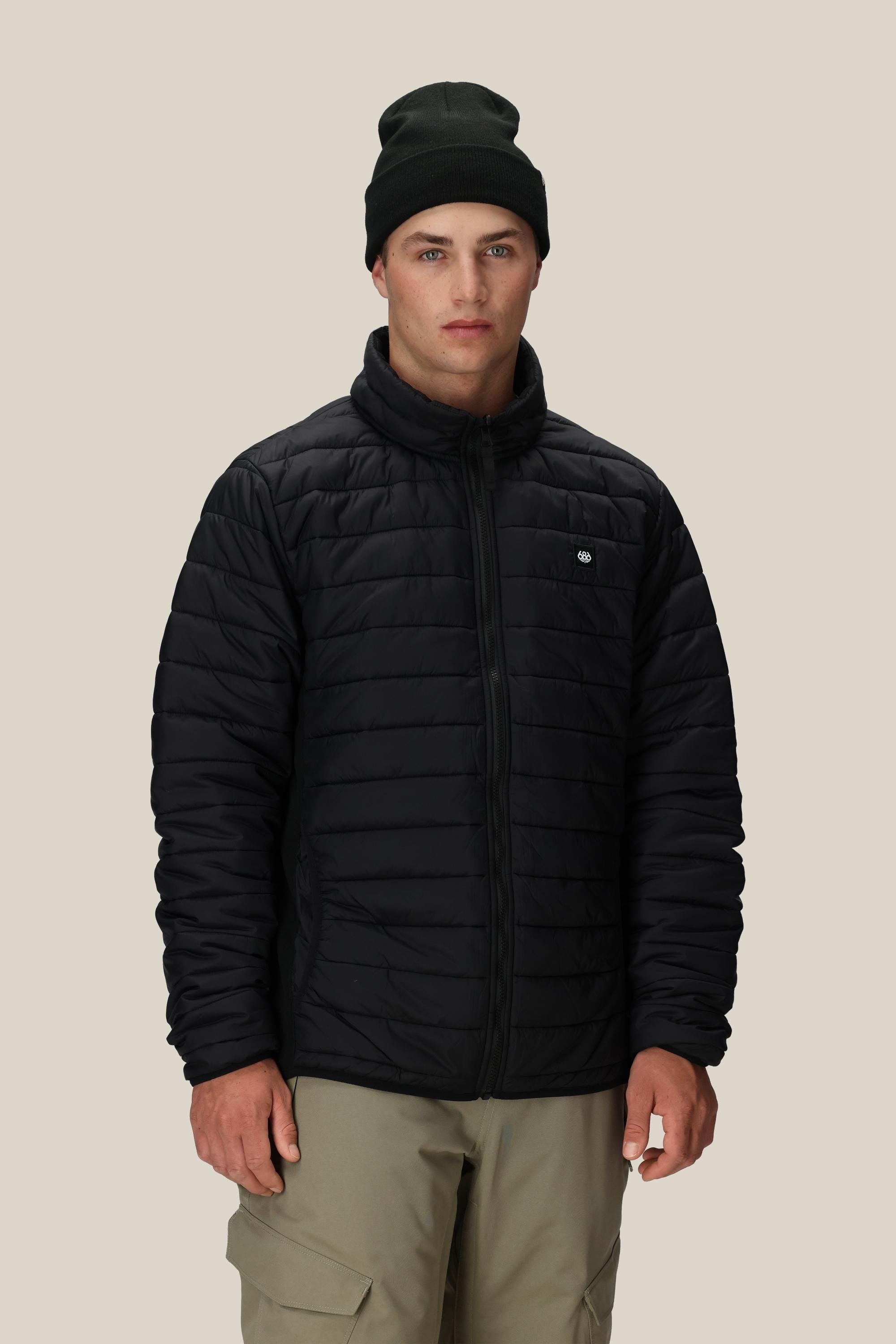 A young man wears the 686 Mens Thermal Puff Jacket by 686 in black, paired with a black beanie and beige pants, standing facing forward against a plain, light beige background.